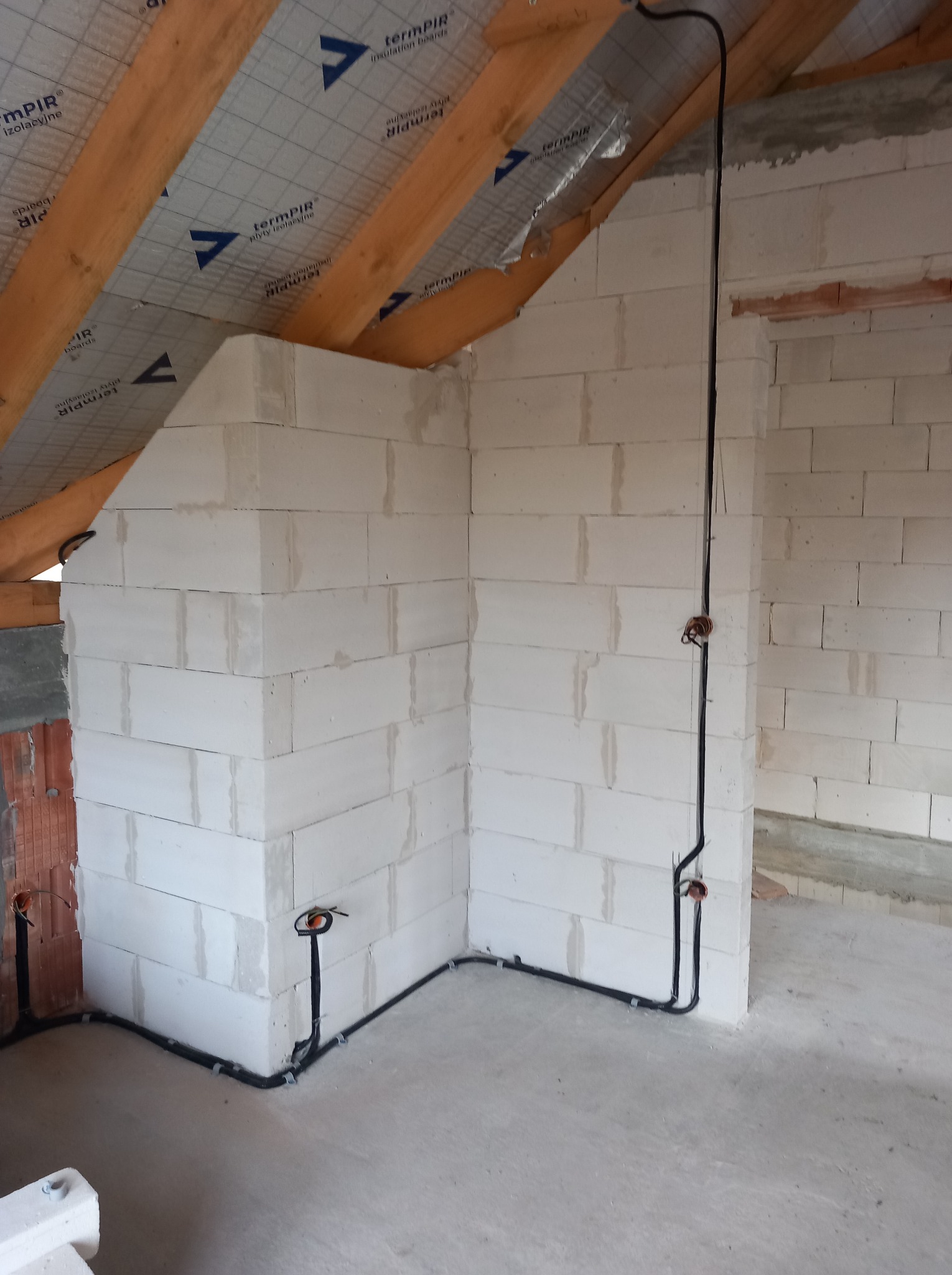 Surowy stan poddasza z widoczną, częściowo rozprowadzoną, czarną instalacją elektryczną na ścianach z białych bloczków. Widoczne drewniane elementy konstrukcji dachu.