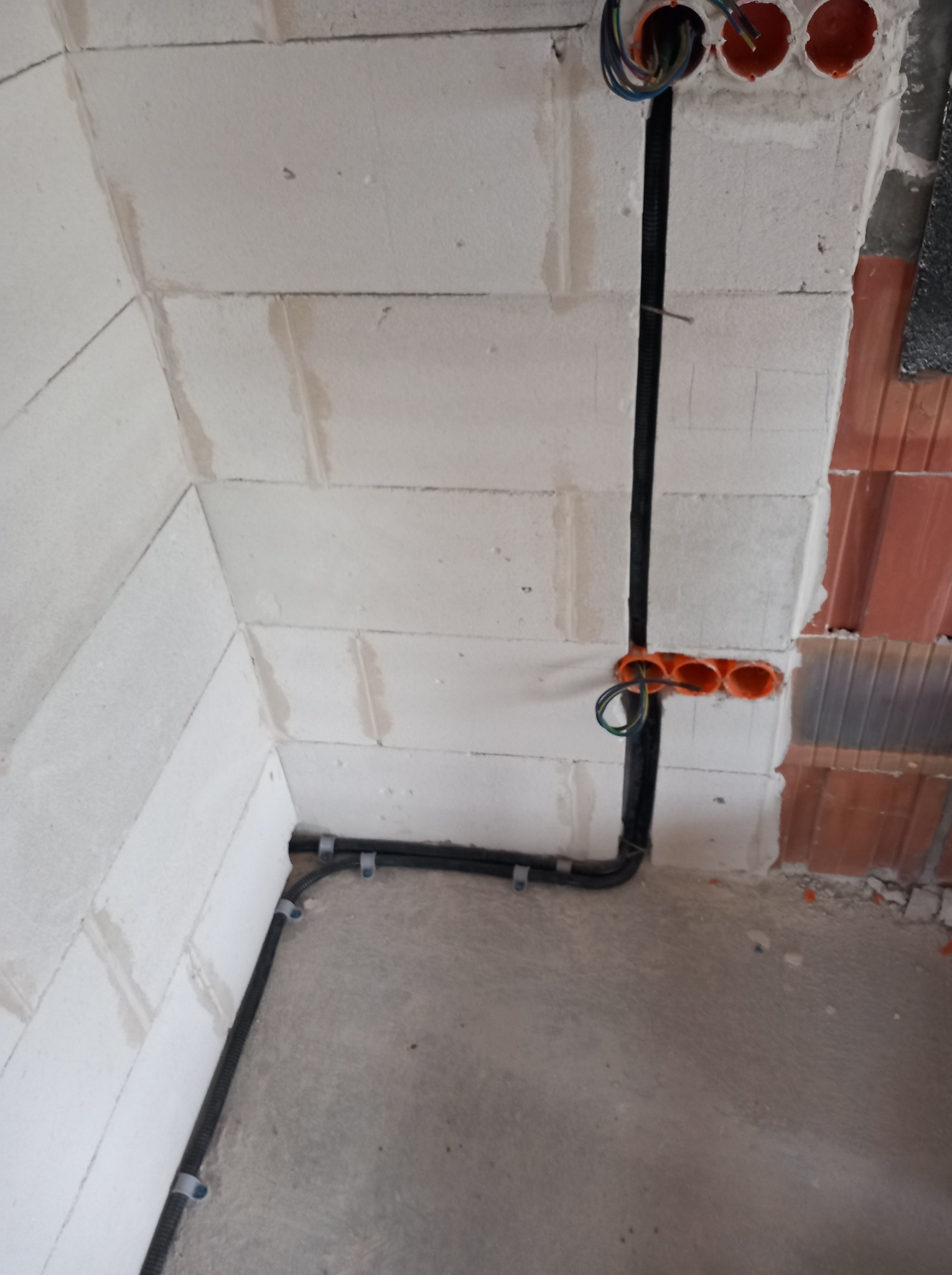 Czarna rura kablowa przymocowana do ściany z betonu komórkowego, z widocznymi pomarańczowymi puszkami instalacyjnymi i przewodami. Surowy stan wykończenia wnętrza.