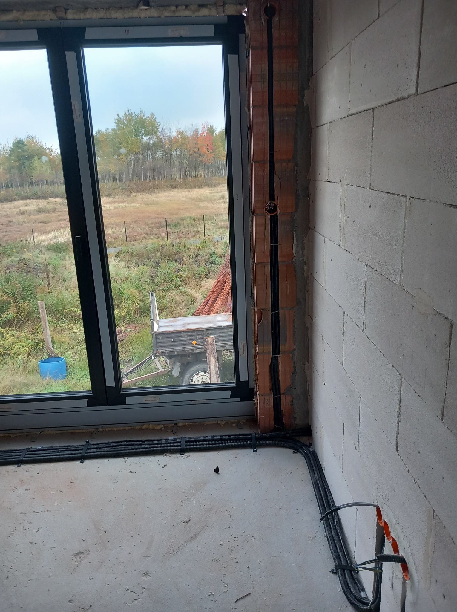 Wewnętrzne kable w peszlach prowadzone wzdłuż ściany z betonu komórkowego, z widocznym oknem i krajobrazem za nim. Instalacja w trakcie realizacji.