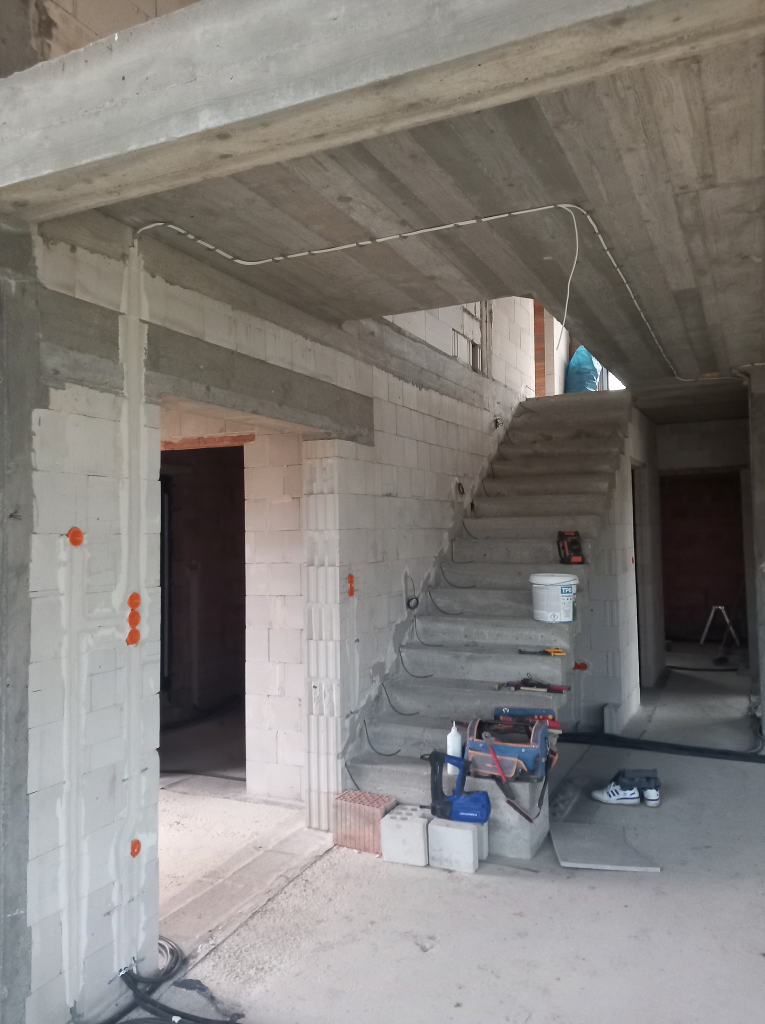 Surowy stan budynku w trakcie prac. Widoczne kable pod sufitem, puszki elektryczne w ścianach, narzędzia na schodach. W tle niezadaszone schody i cegły.