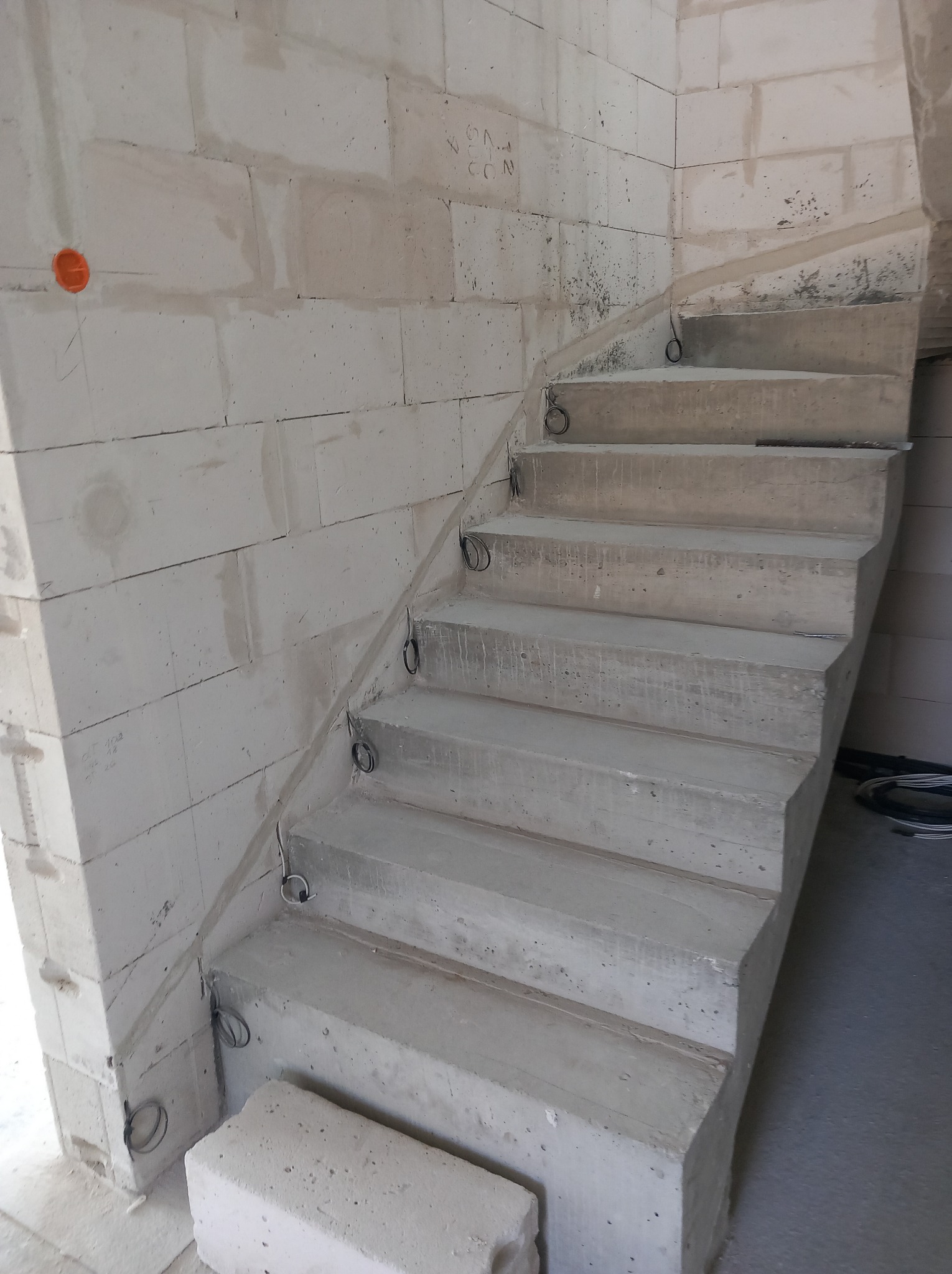 Surowe betonowe schody spiralne w surowym stanie, z widocznymi przewodami elektrycznymi w ścianie z białych bloczków. Pomarańczowa puszka elektryczna na ścianie.