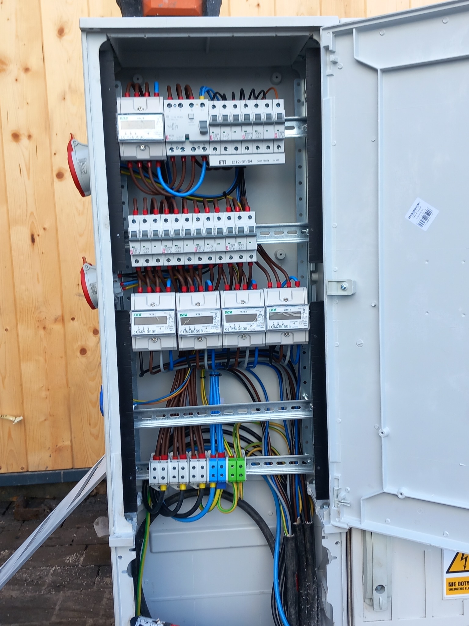 Otwarta skrzynka elektryczna z widocznymi bezpiecznikami, przewodami w kolorach: niebieskim, brązowym, czarnym i zielono-żółtym. Obok drewniana ściana i ostrzeżenie o napięciu.