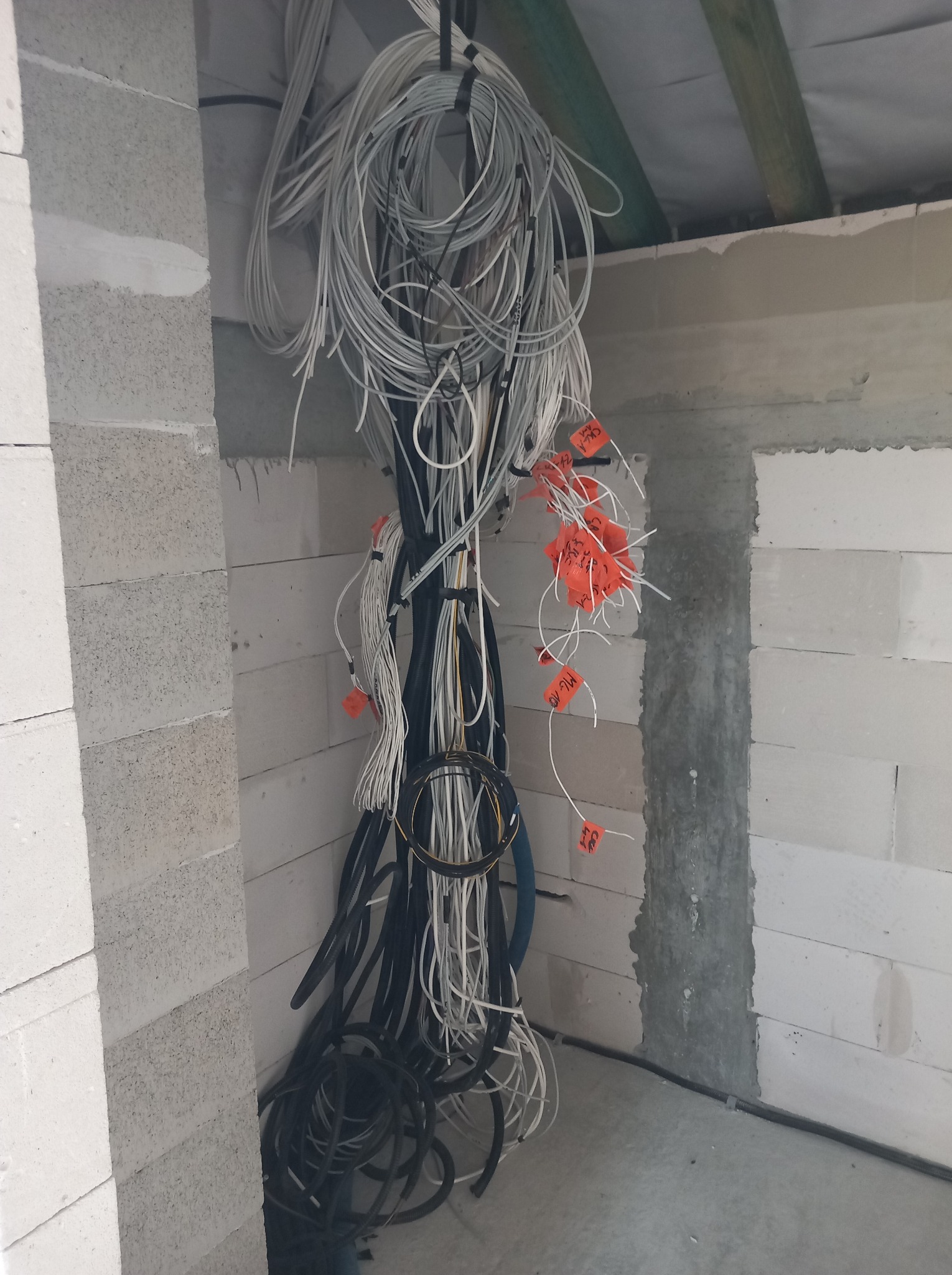 Wiązka przewodów elektrycznych z pomarańczowymi etykietami, zwisająca ze stropu w surowym wnętrzu z białych bloczków. Widoczne kable w różnych kolorach i grubościach.