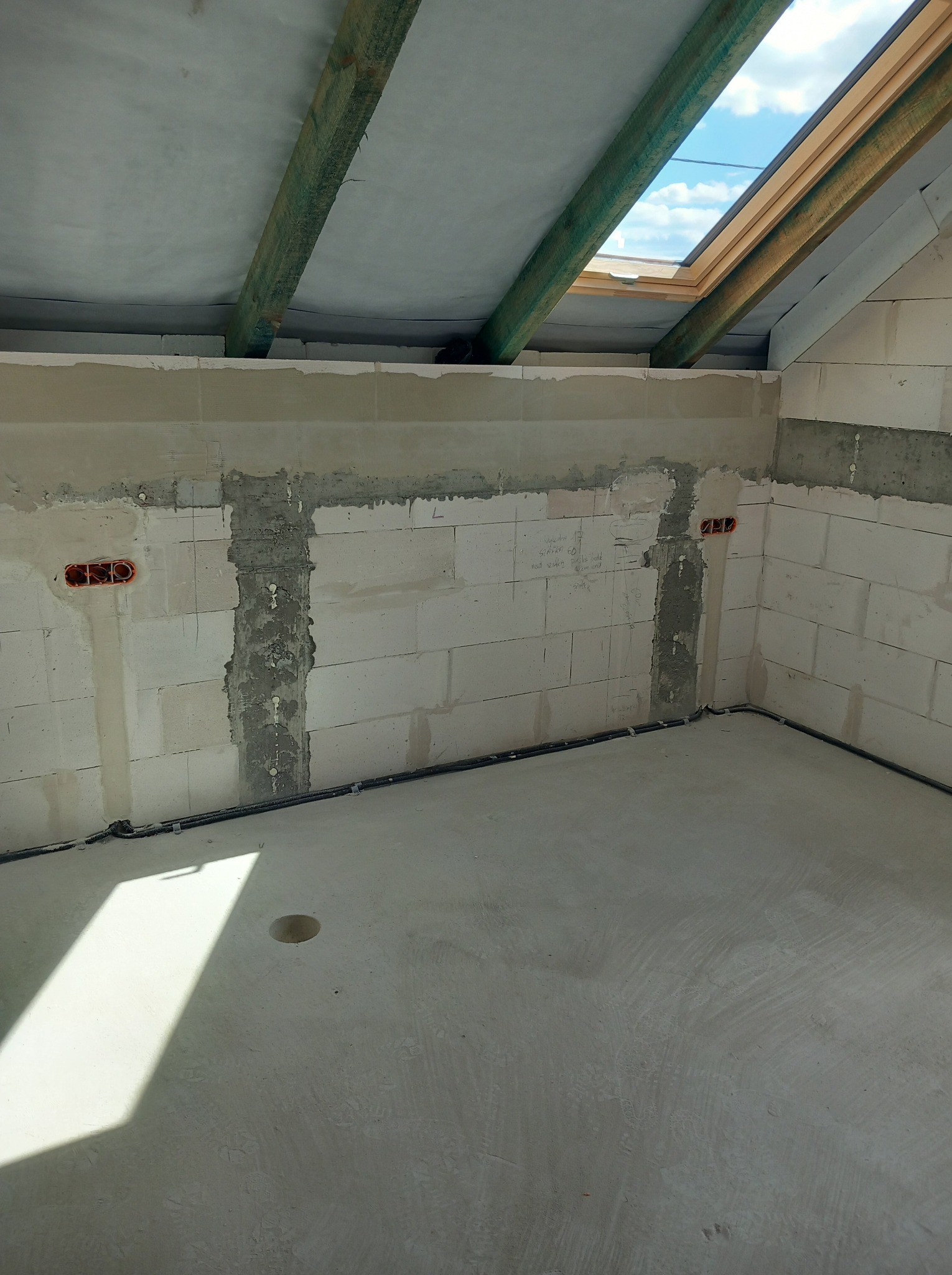 Surowy stan poddasza z oknem dachowym, widoczne kable elektryczne w ścianach z betonu komórkowego i wylewka podłogowa z otworem. Etap prac instalacyjnych.