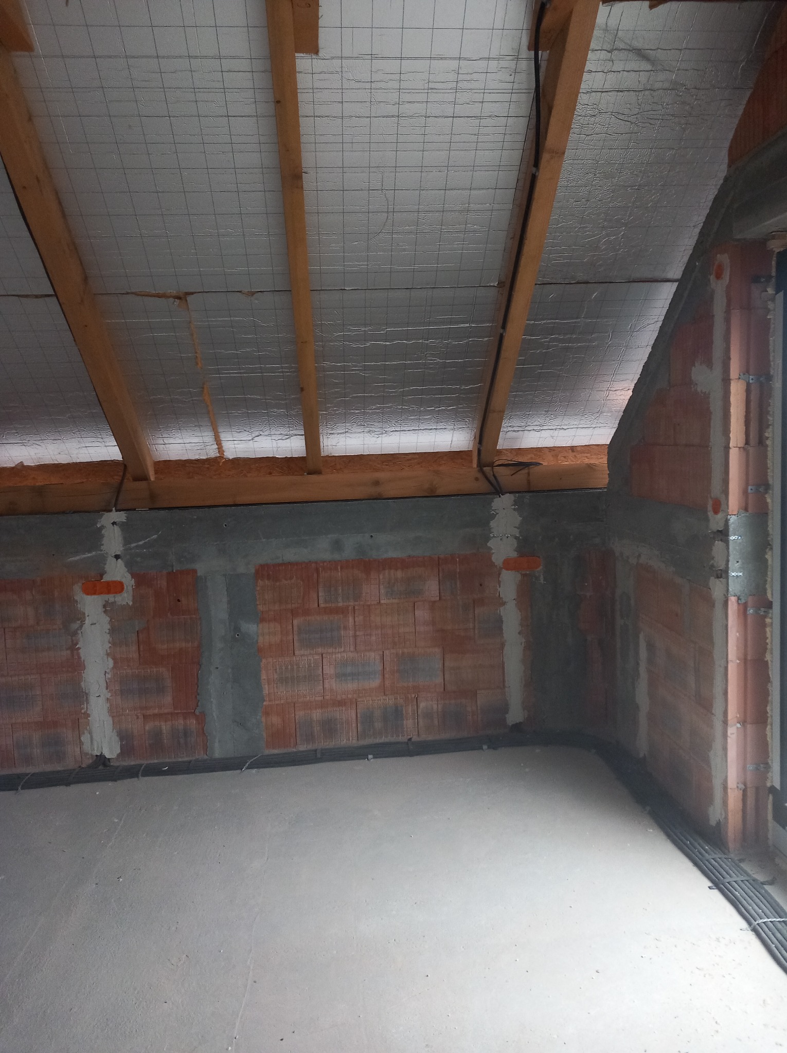 Surowy stan poddasza z widoczną izolacją termiczną pod dachem, murem z cegły i przewodami elektrycznymi przy podłodze. Wnętrze w trakcie prac wykończeniowych.