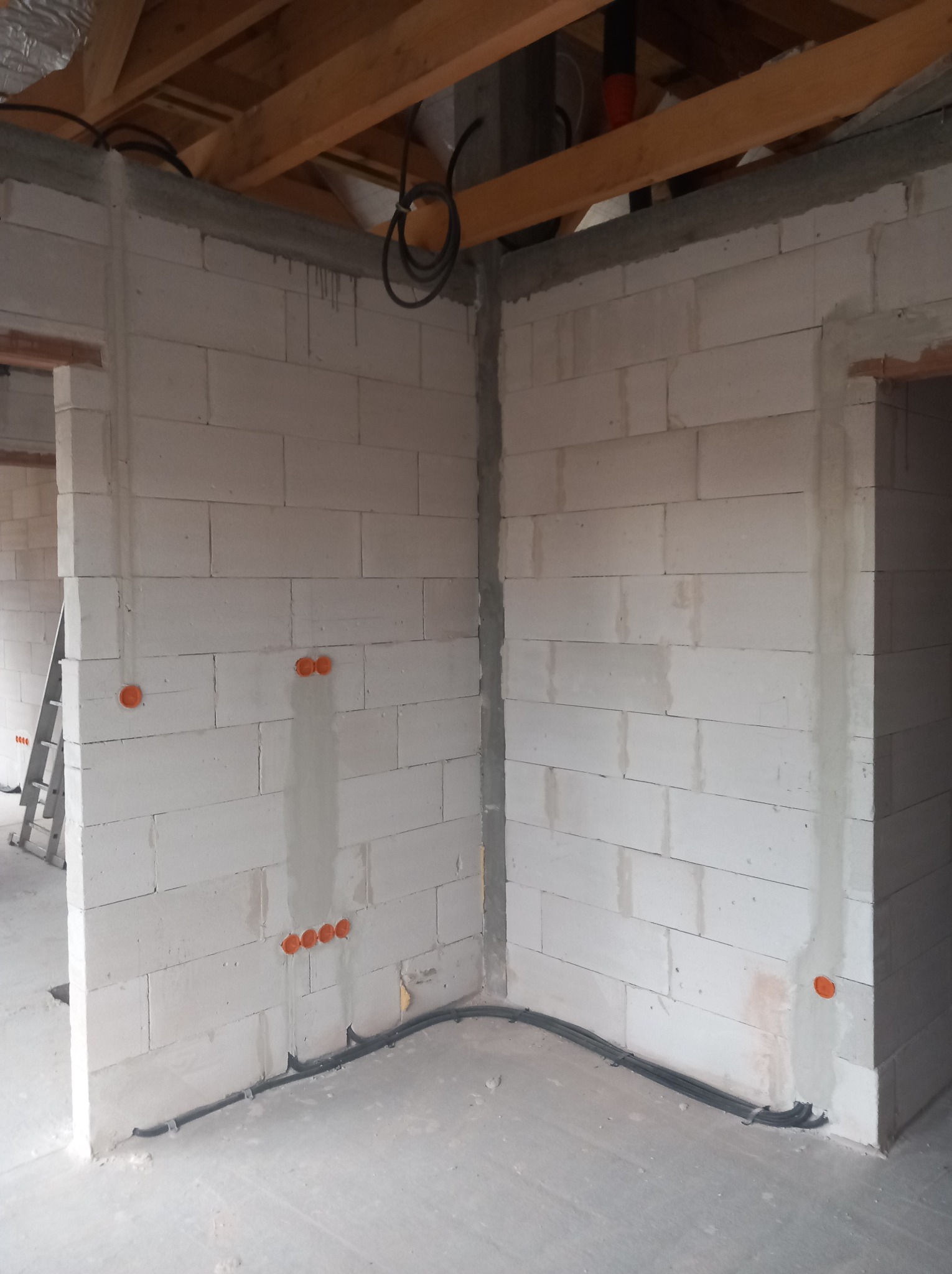 Wnętrze w trakcie budowy z widocznymi przewodami elektrycznymi ułożonymi wzdłuż ściany z betonu komórkowego i puszkami instalacyjnymi w kolorze pomarańczowym.