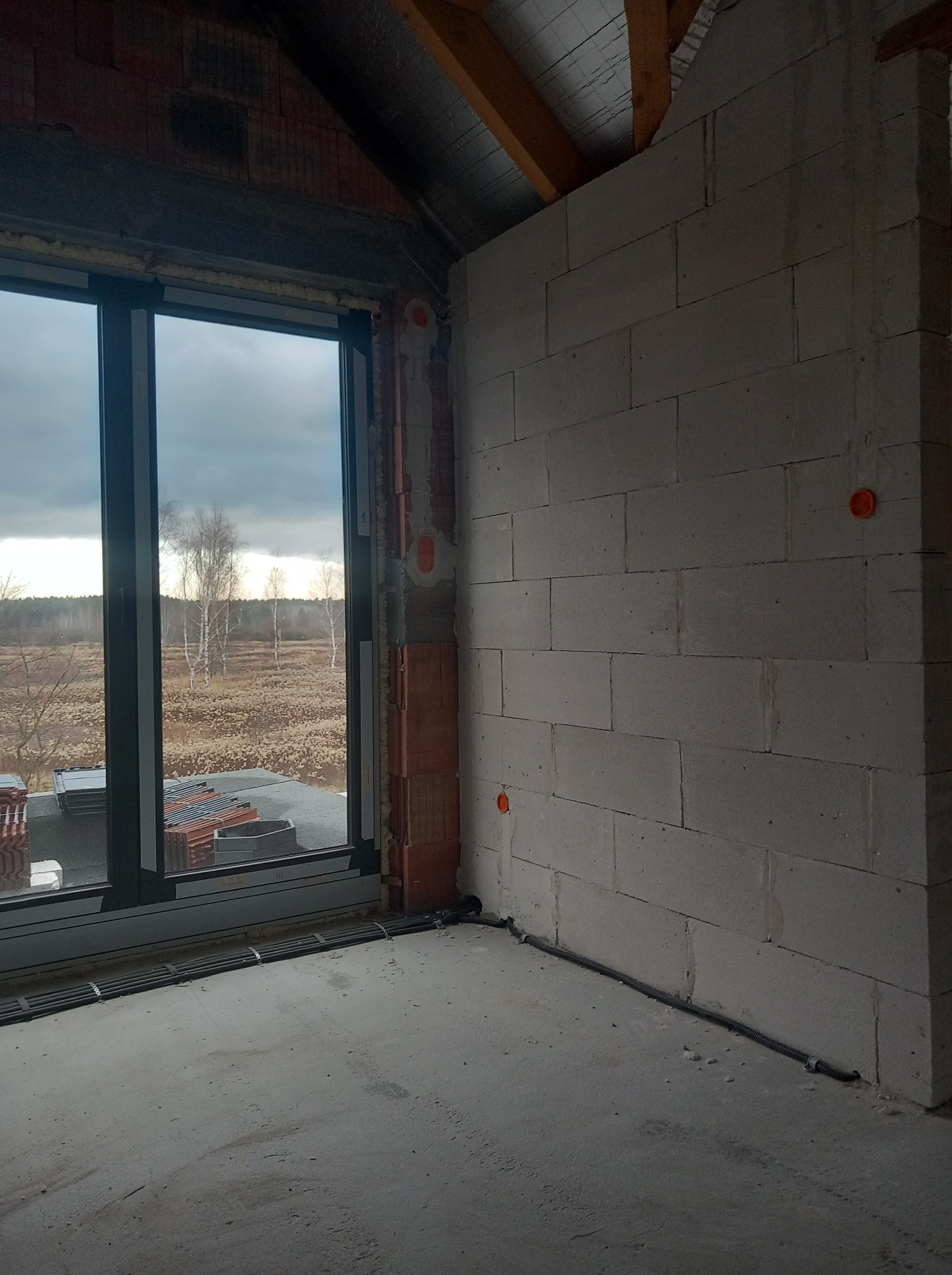 Wnętrze w stanie surowym w Jarocinie: widoczne przewody w ścianie z betonu komórkowego i peszle na podłodze, okno z widokiem na zewnątrz. Instalacja w trakcie.