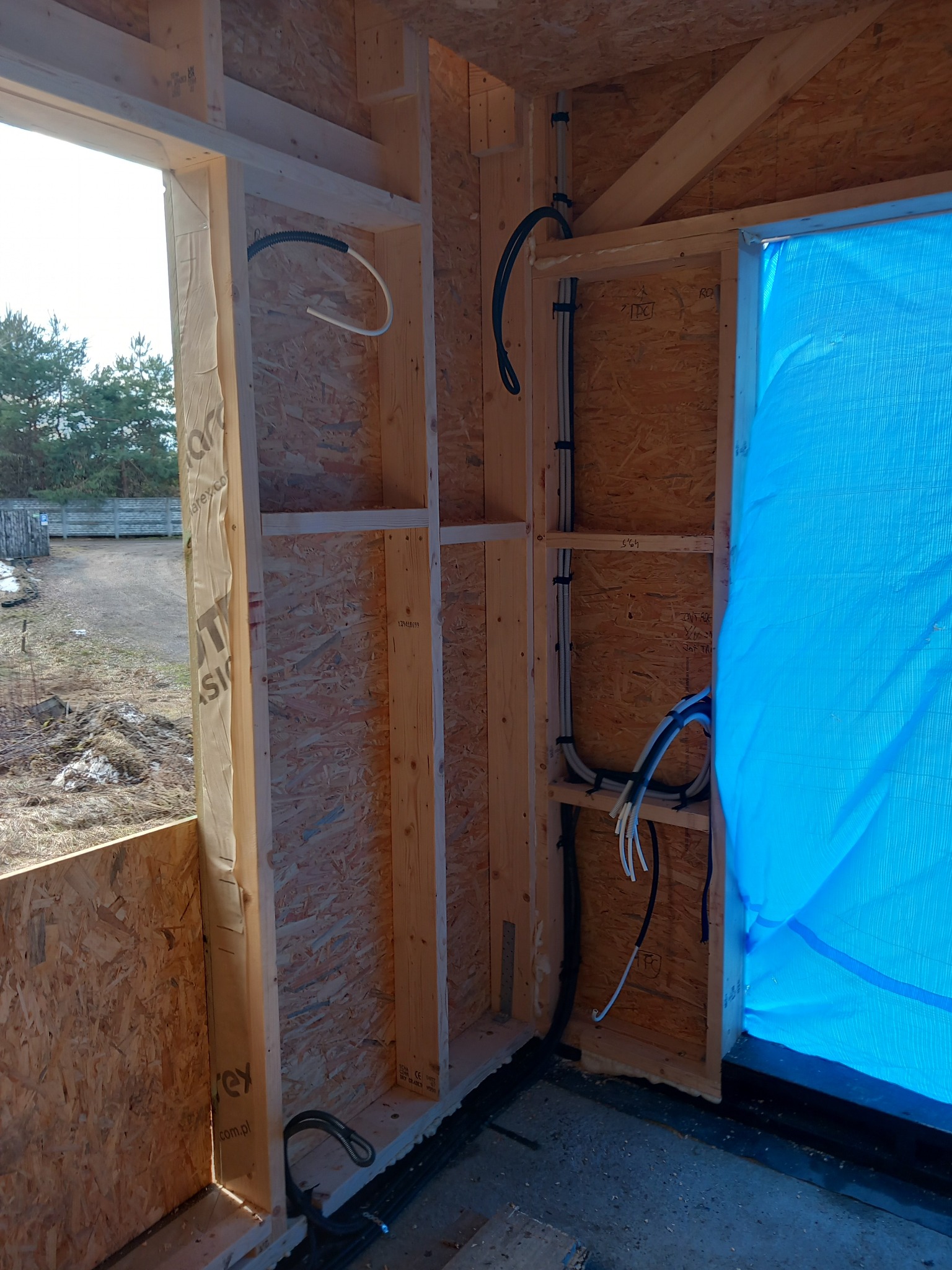 Wnętrze szkieletowej konstrukcji domu w trakcie budowy z widocznymi przewodami elektrycznymi przymocowanymi do ściany z płyty OSB. Częściowo widoczne okno i niebieska folia.