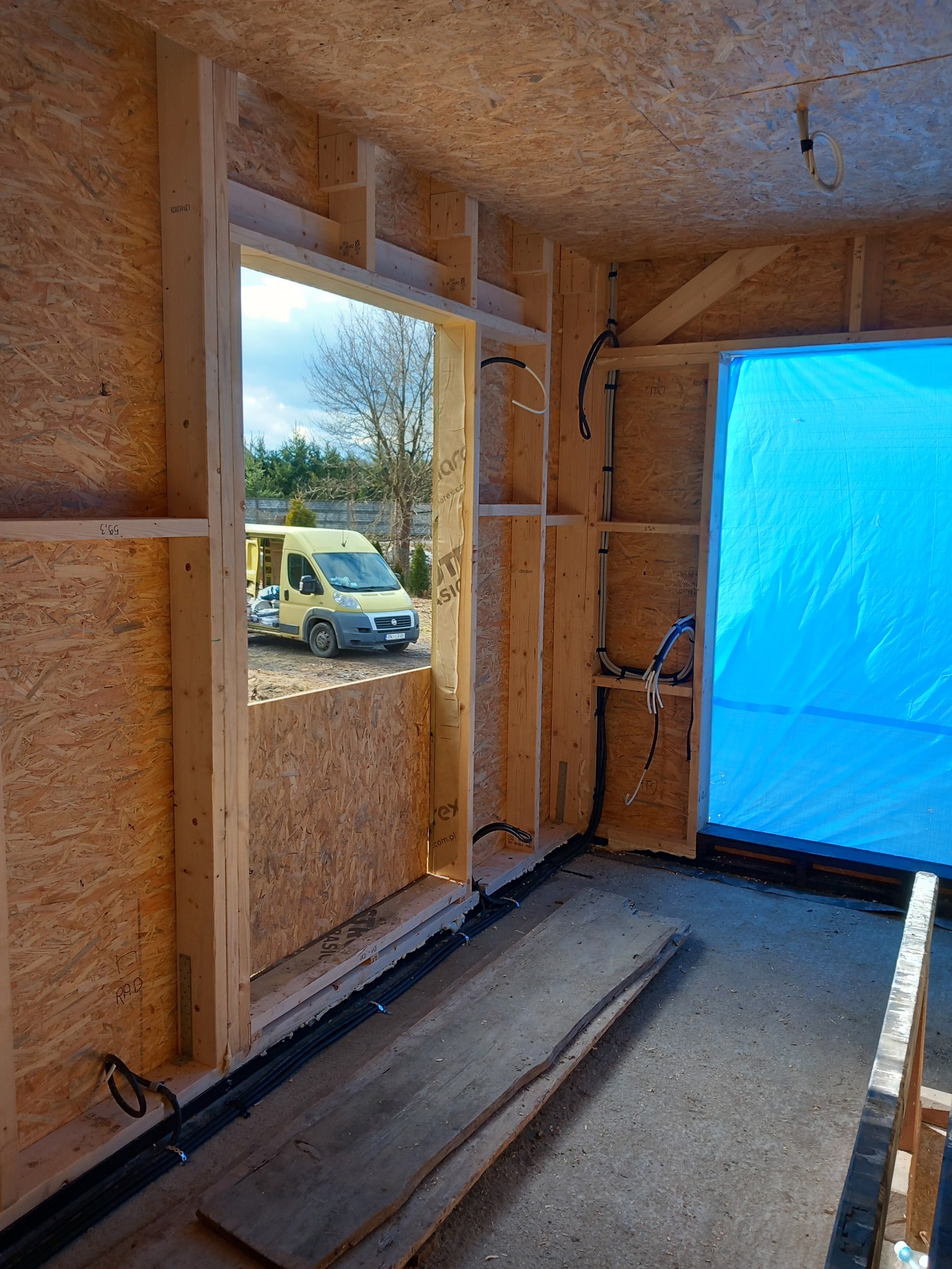 Wnętrze szkieletowej konstrukcji domu w budowie z widocznymi przewodami elektrycznymi w ścianach i podłodze. Na zewnątrz żółty bus.