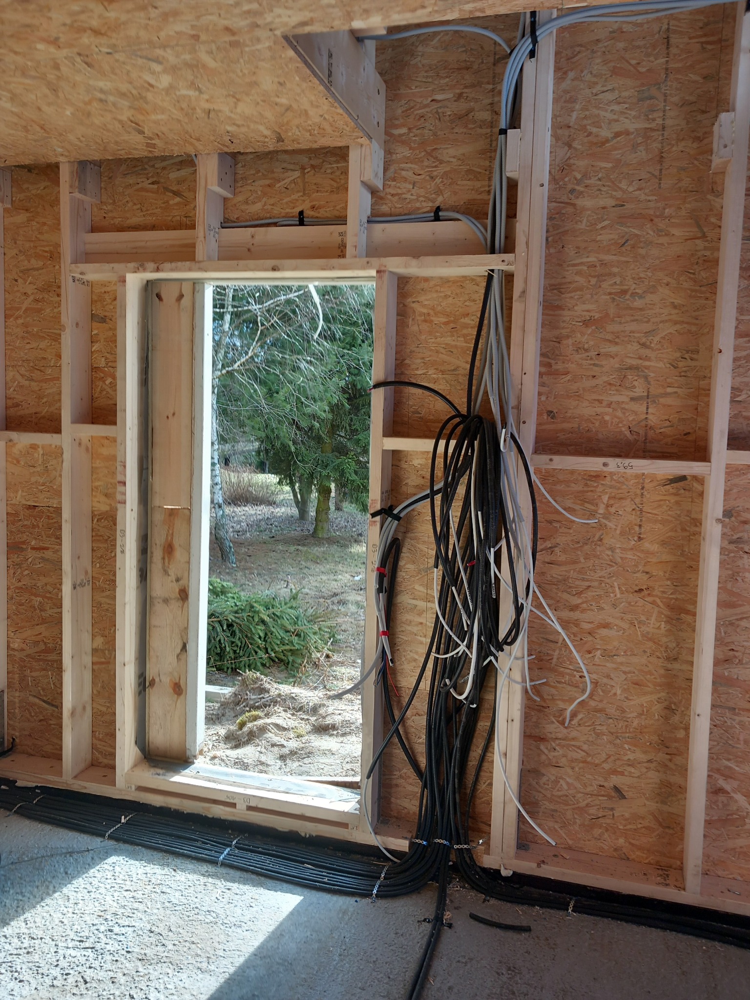 Instalacja elektryczna w surowym budynku: kable prowadzone w ścianach z płyty OSB, widoczne przewody pod podłogą i przez otwór okienny. Etap prac przed wykończeniem.