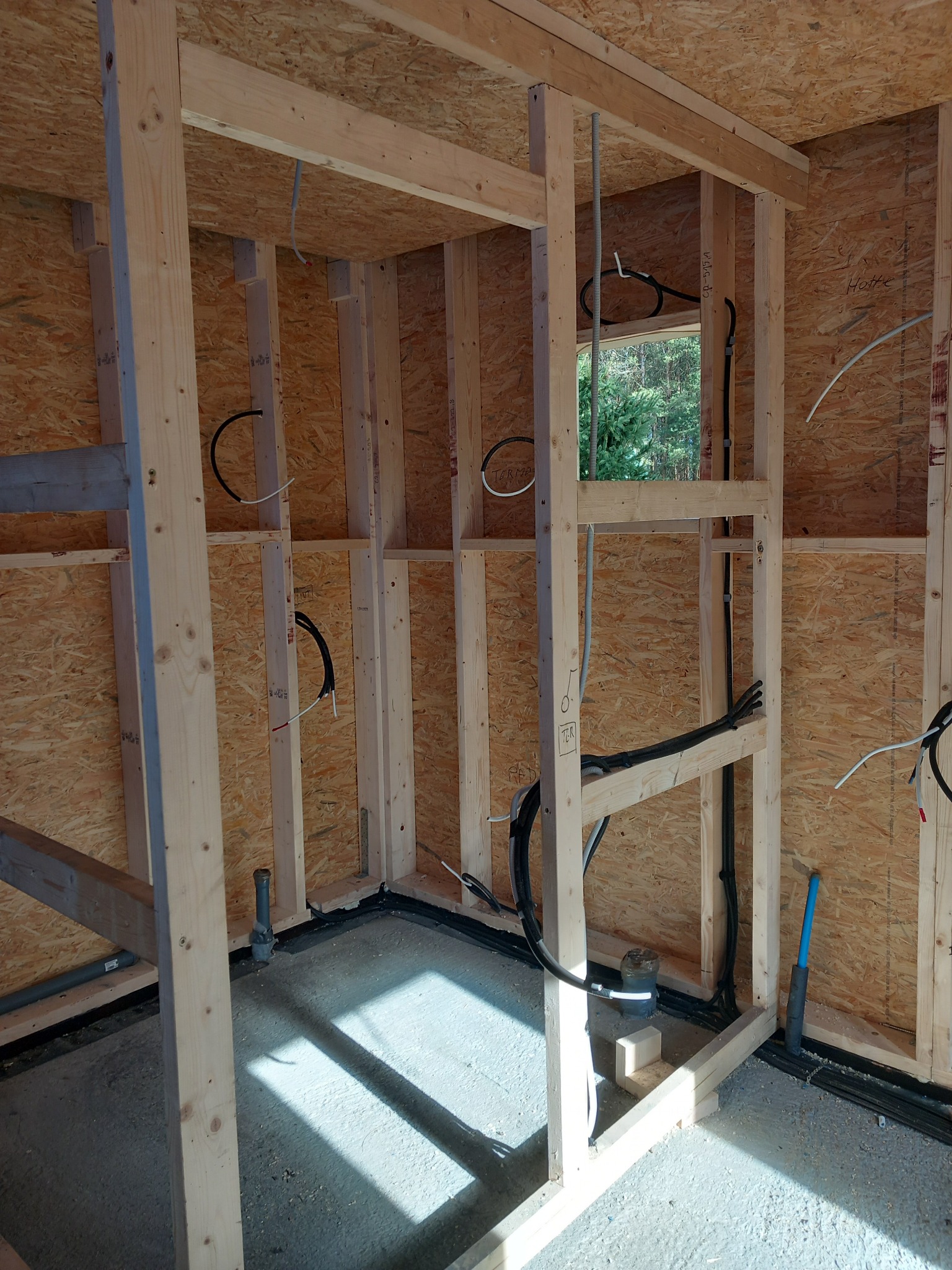 Surowy szkielet drewniany z widocznymi przewodami i rurami instalacji wewnątrz ścian. W tle okno z widokiem na zieleń. Widać etap prac instalacyjnych w nowym budynku.