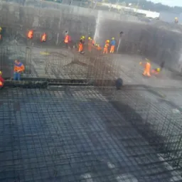 Zbrojenie terenu budowy z pracownikami w odblaskowych strojach, widok z góry. Siatka z prętów zbrojeniowych na czarnej folii. Prace zbrojarskie w toku.