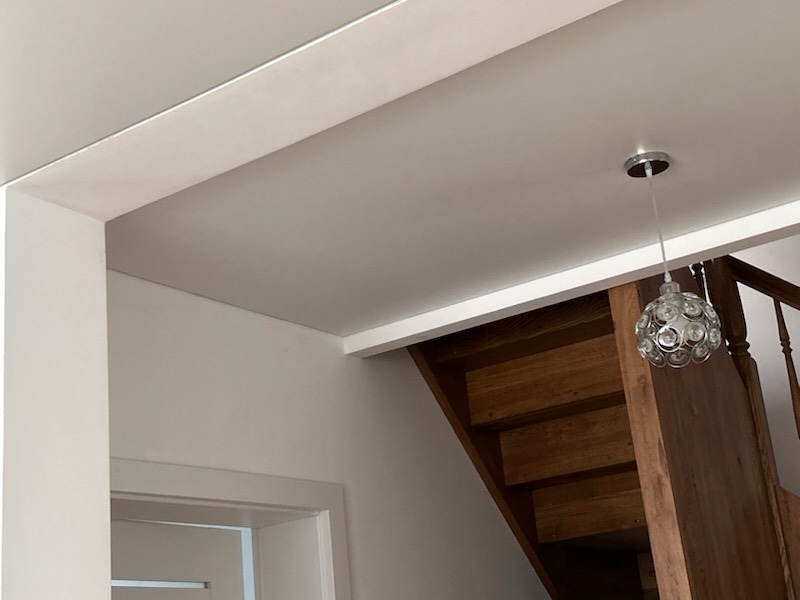 Jasny, gładki sufit w nowym domu, z widoczną lampą wiszącą w kształcie kuli i fragmentem drewnianych schodów w tle.