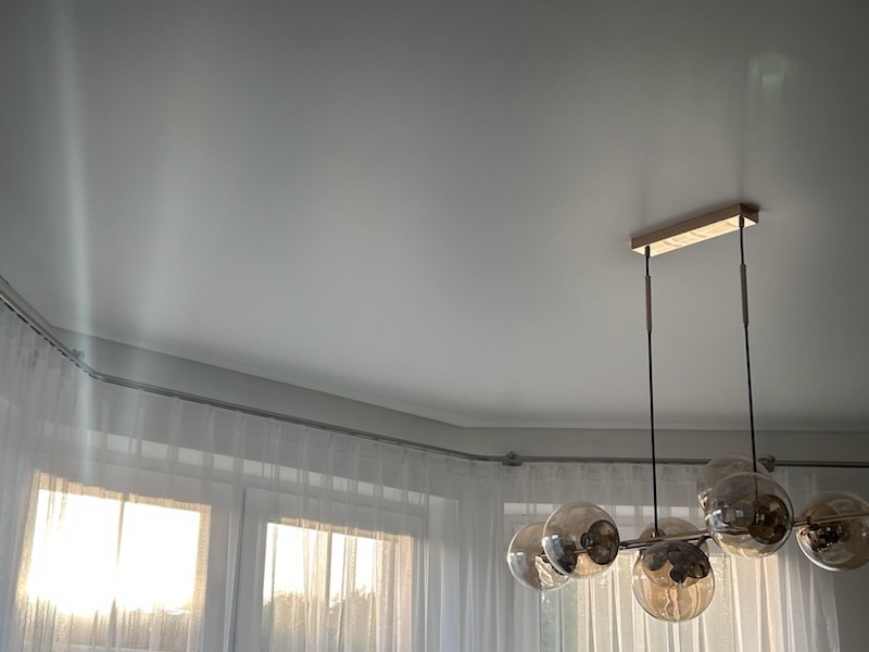 Gładki, biały sufit z połyskiem, oświetlony nowoczesną lampą wiszącą z kulistymi kloszami; widoczne okno z firankami.