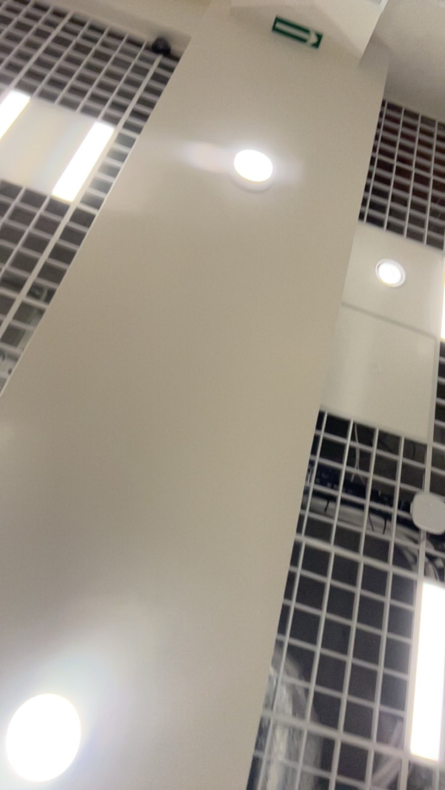 Biały sufit z podwieszanymi panelami rastrowymi i okrągłymi lampami LED, widok z dołu pod kątem.