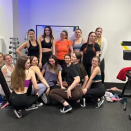 Grupa kobiet w strojach sportowych pozuje w siłowni, widoczny sprzęt do ćwiczeń, tablica z zapisanymi notatkami i butelki z napojami.