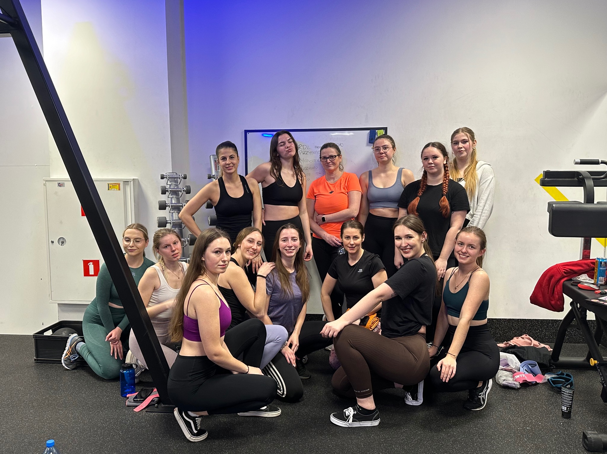 Grupa kilkunastu kobiet w strojach sportowych pozuje w siłowni na tle białej tablicy i sprzętu do ćwiczeń.