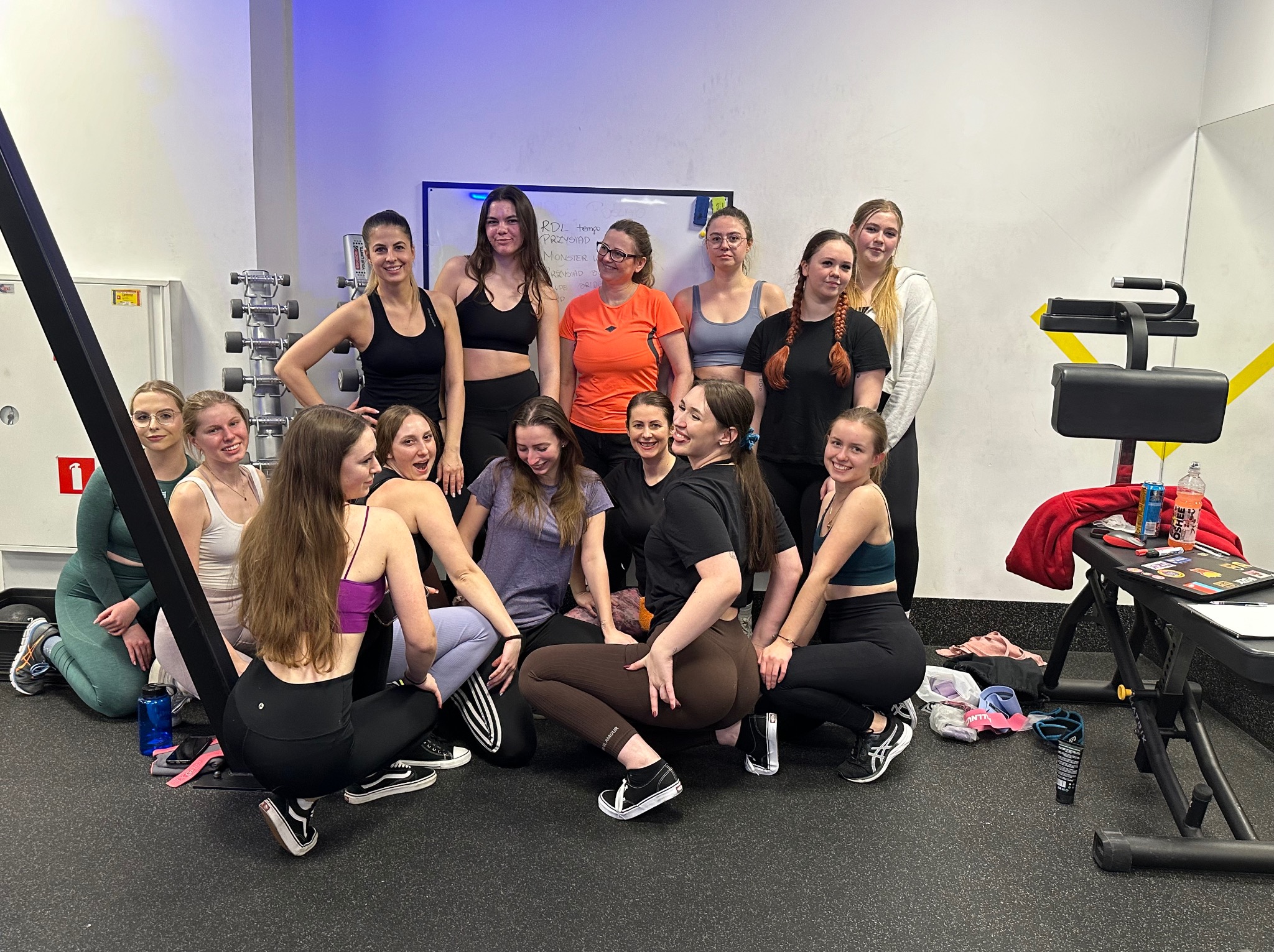 Grupa kobiet w strojach sportowych pozuje w siłowni, widoczny sprzęt do ćwiczeń, tablica z zapisanymi notatkami i butelki z napojami.