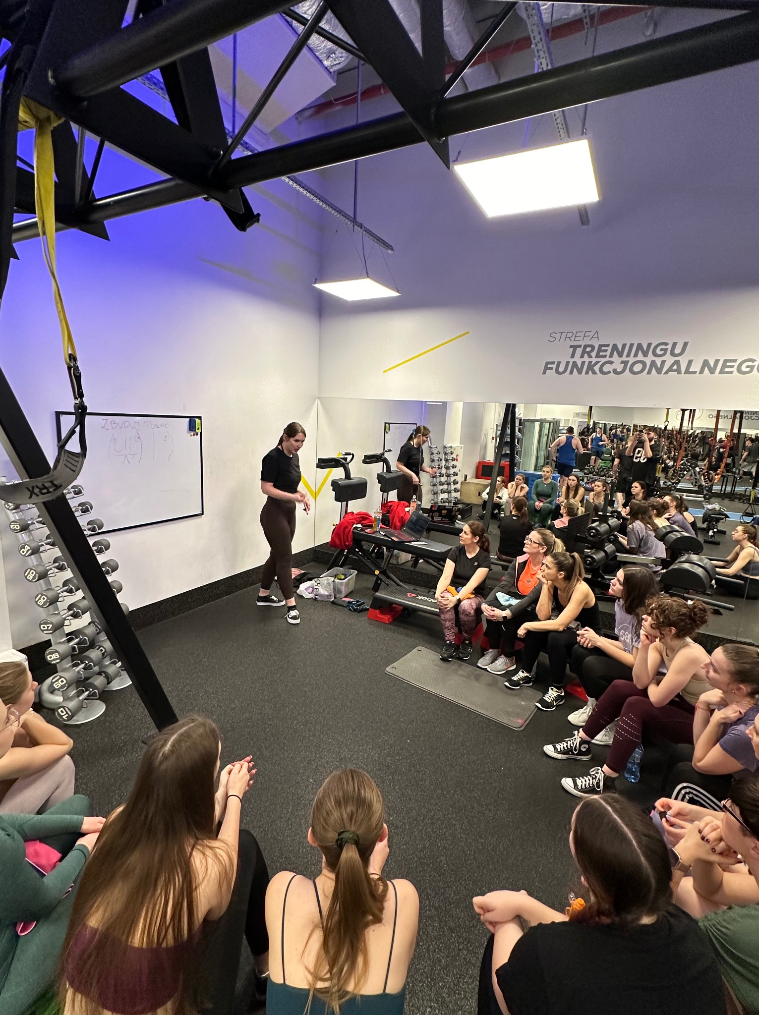 Instruktorka prowadzi wykład o treningu funkcjonalnym dla grupy kobiet w siłowni, na tablicy rysunek z opisem ćwiczeń.