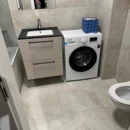 OK-Bud Oleh Kopytko - Ustawiona pralka Samsung obok umywalki z szafką i toalety w łazience wykończonej płytkami imitującymi marmur, z odbiciem osoby w lustrze.