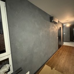 OK-Bud Oleh Kopytko - Wnętrze po wykonaniu gładzi dekoracyjnej w odcieniach szarości, widoczny fragment okna z parapetem, drewniana podłoga i czarne drzwi w głębi pomieszczenia.