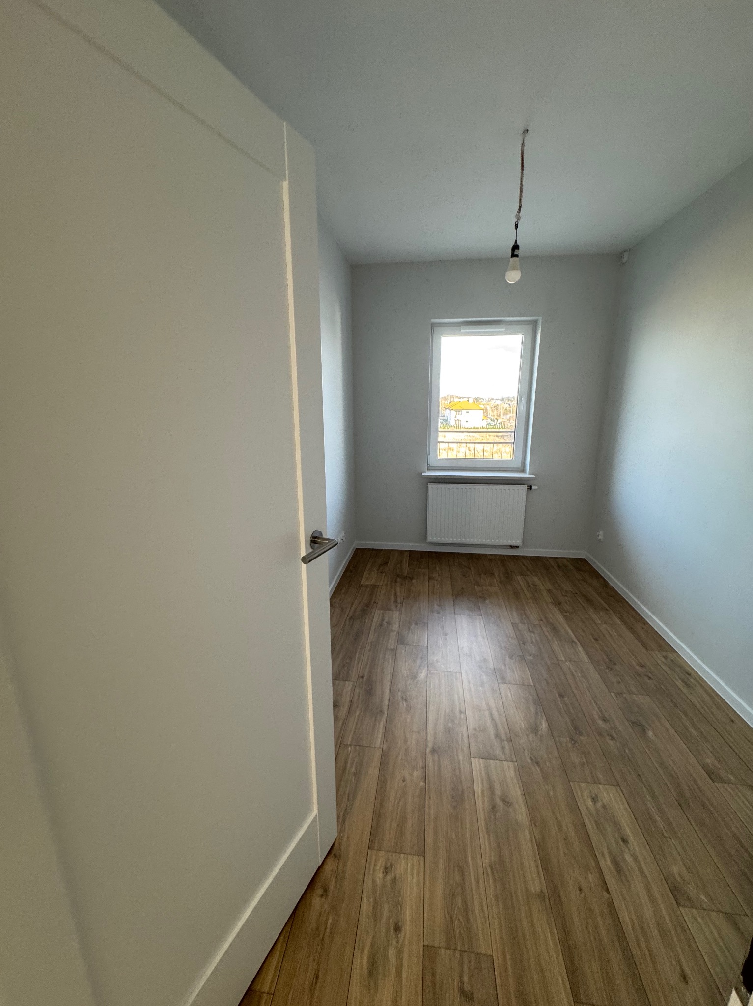 Pusty pokój z drewnianą podłogą i świeżo pomalowanymi ścianami na biało, widoczne okno z grzejnikiem, drzwi wejściowe z klamką i wisząca żarówka bez oprawy.