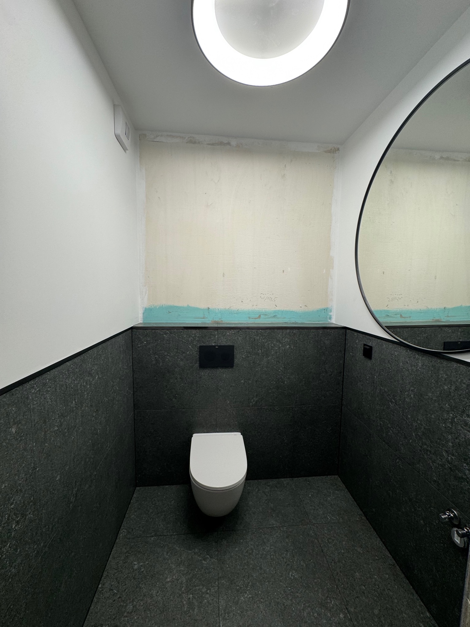 Częściowo wykończona łazienka z czarnymi płytkami na ścianach i podłodze, białą wiszącą toaletą i okrągłym lustrem. Na ścianie widoczne ślady po wcześniejszych pracach, z fragmentami niepokrytej...