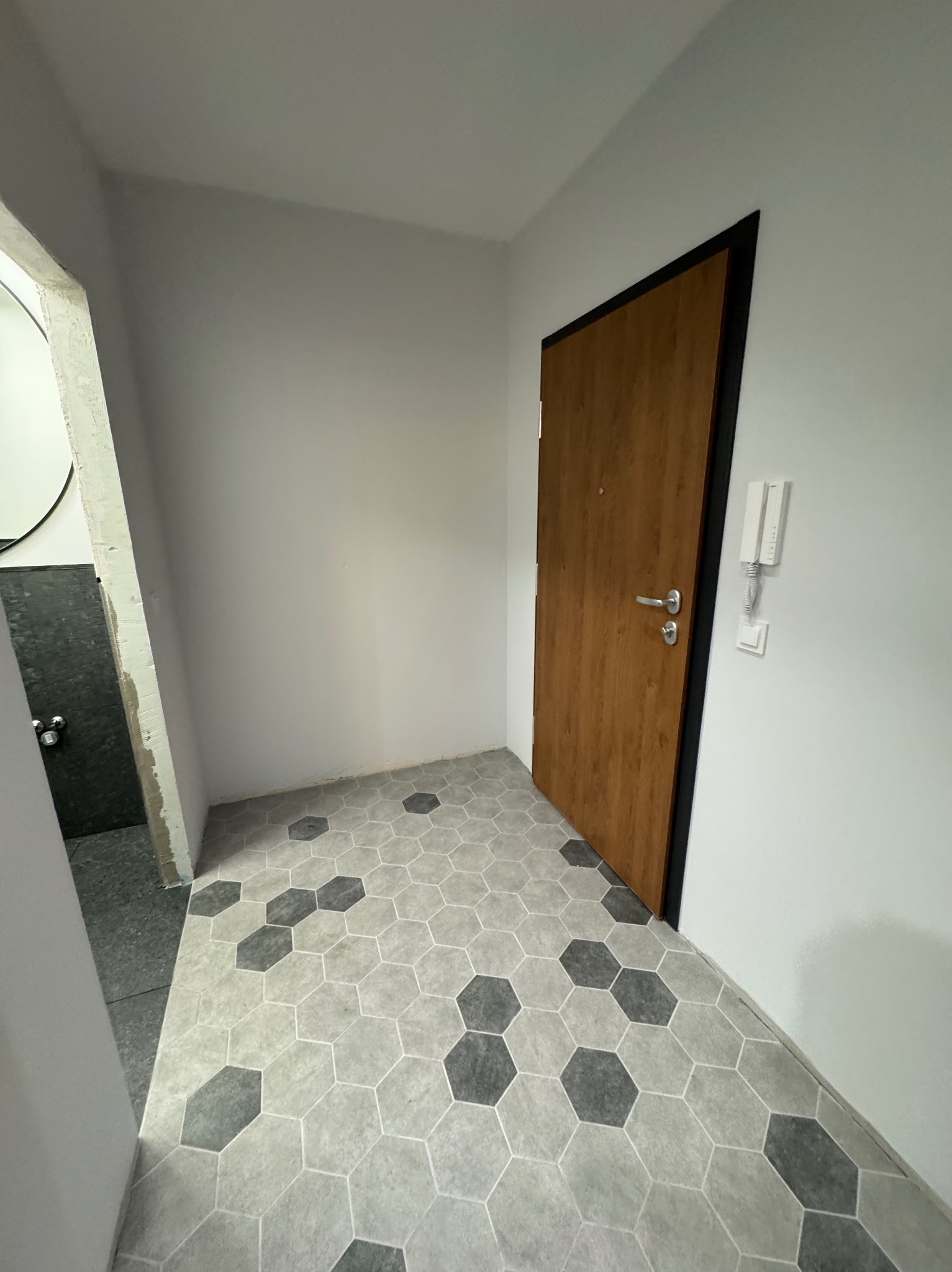 Przedpokój po remoncie z szarą heksagonalną podłogą, drewnianymi drzwiami z czarną ościeżnicą i białymi ścianami z domofonem.