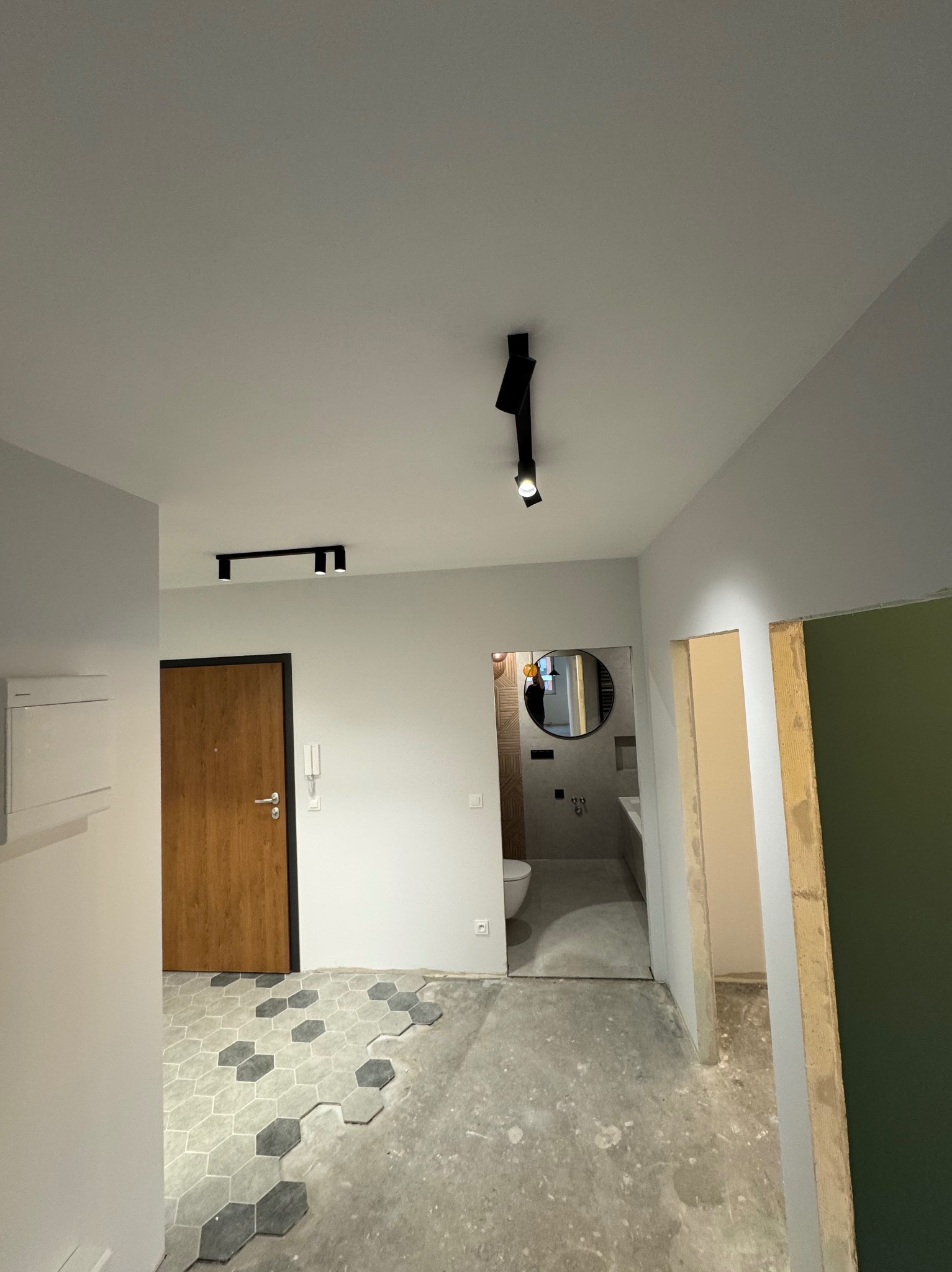 Wnętrze mieszkania w trakcie remontu: położone częściowo heksagonalne płytki na podłodze, widoczna łazienka z okrągłym lustrem, surowa ściana przygotowana do malowania, nowoczesne czarne...