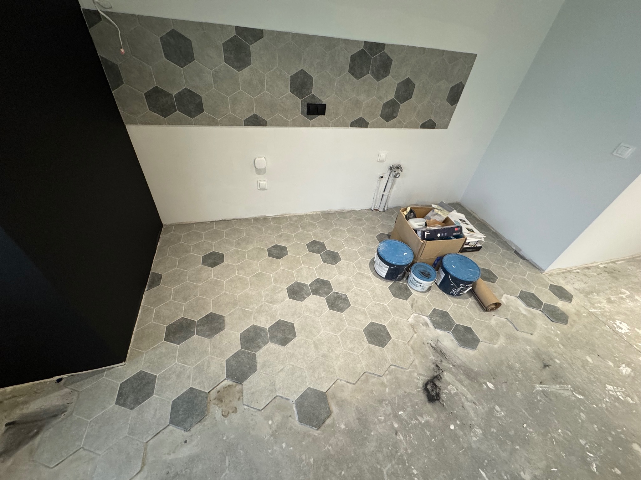 Łazienka w trakcie remontu: położone szare heksagonalne płytki podłogowe i częściowo na ścianie, widoczne puszki z farbą, karton i rury hydrauliczne.