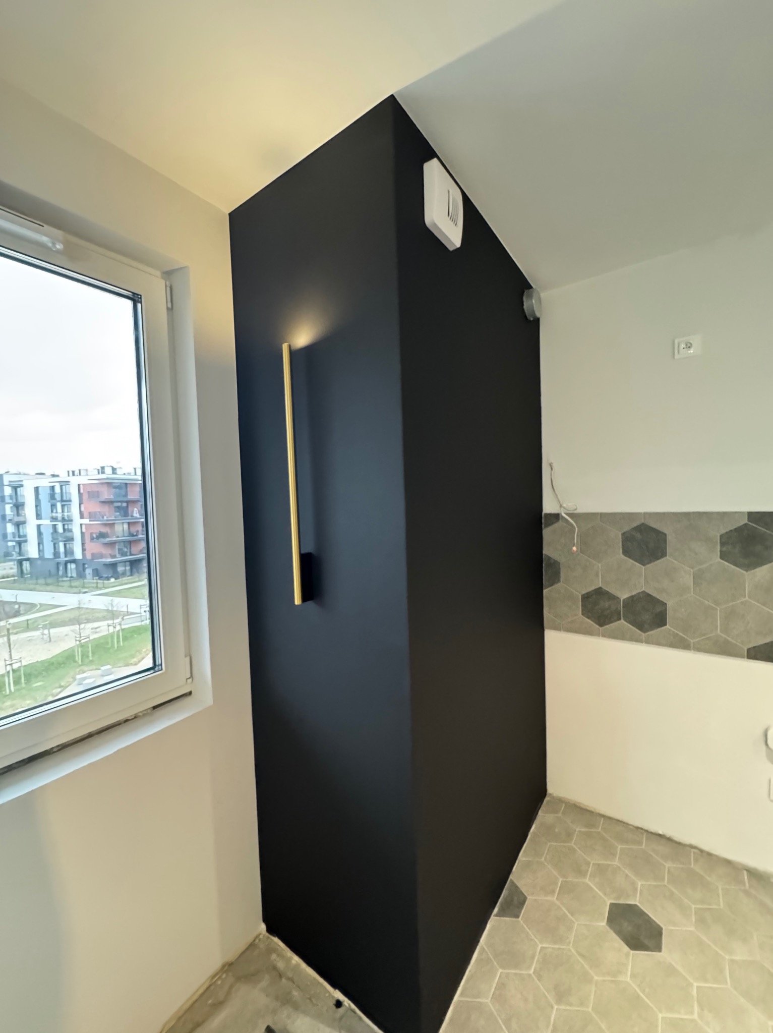 Pomieszczenie po remoncie: czarna ściana z podłużnym złotym kinkietem, heksagonalna podłoga, okno z widokiem na budynki.