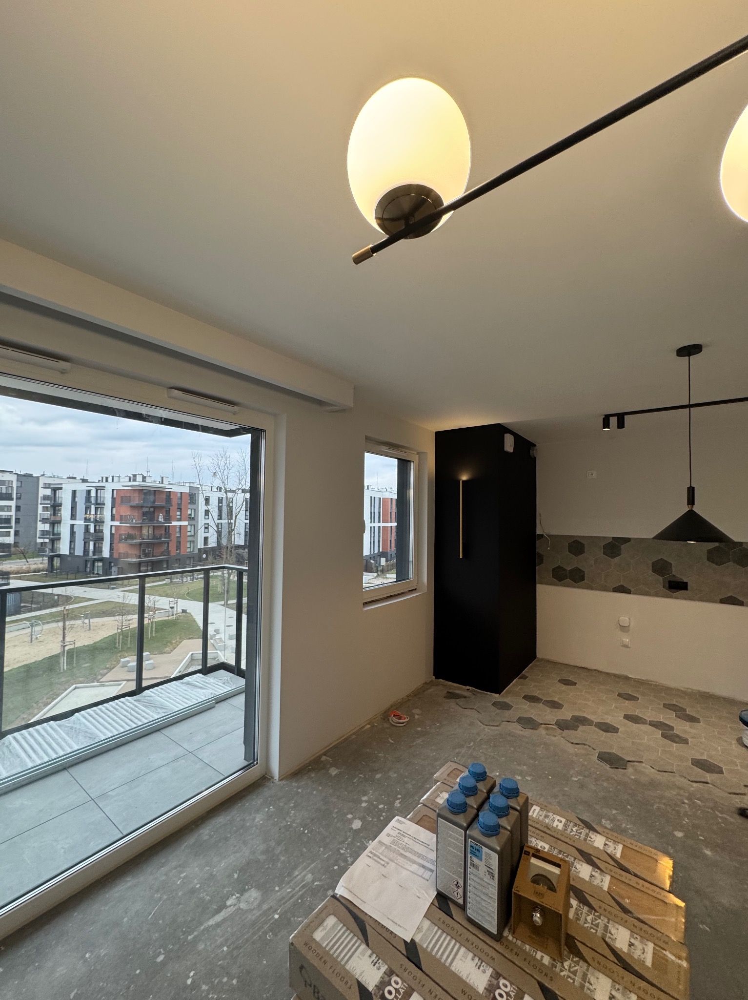 Wnętrze mieszkania w trakcie układania heksagonalnych płytek podłogowych, widoczne opakowania paneli i preparatów do podłóg, nowoczesne oświetlenie sufitowe, duże okno balkonowe z widokiem na osiedle.