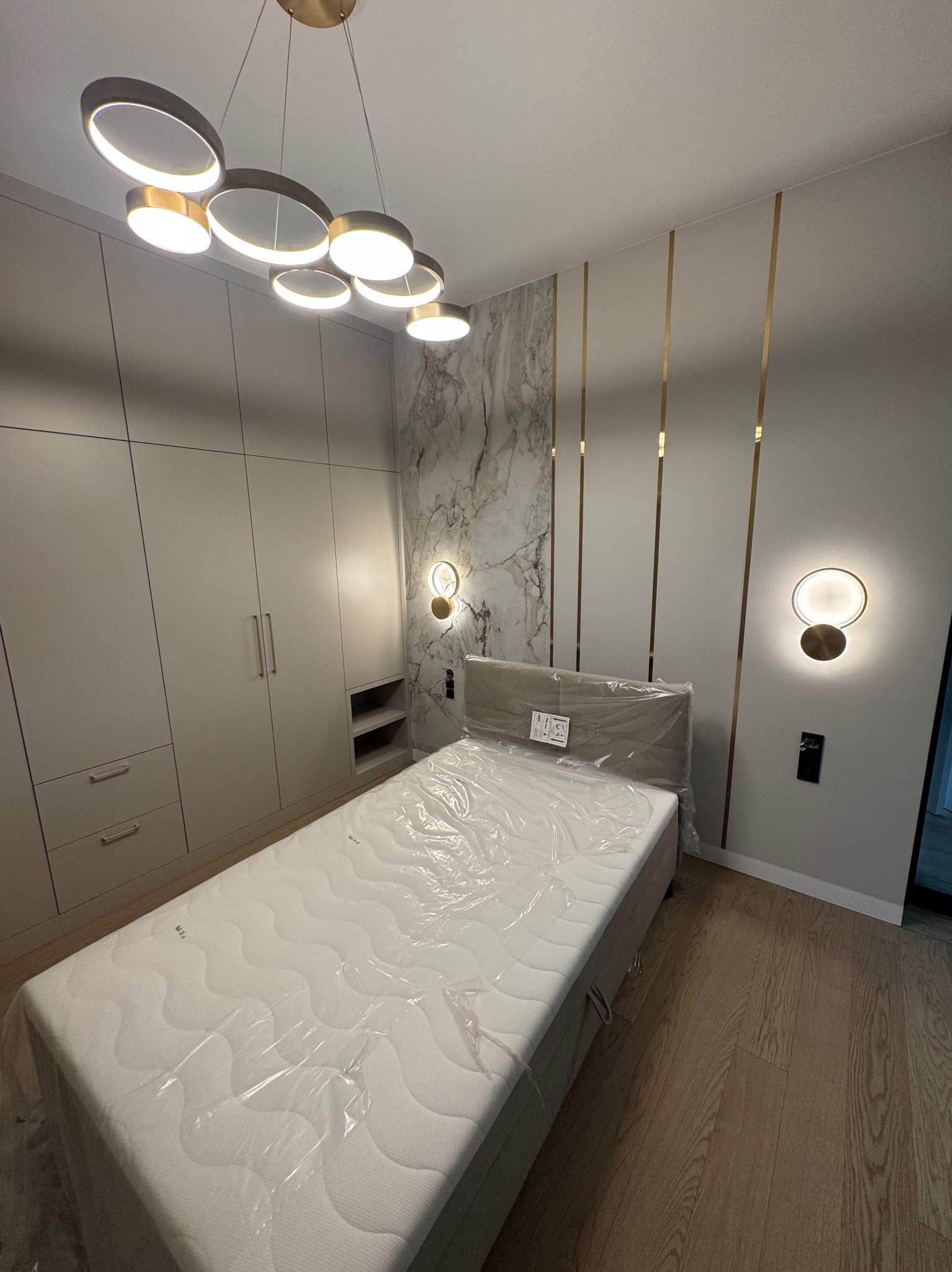 Nowoczesna sypialnia z łóżkiem przykrytym folią ochronną, zabudowaną szafą z szarymi frontami, marmurową ścianą akcentową i oświetleniem LED w postaci okręgów i kinkietów.