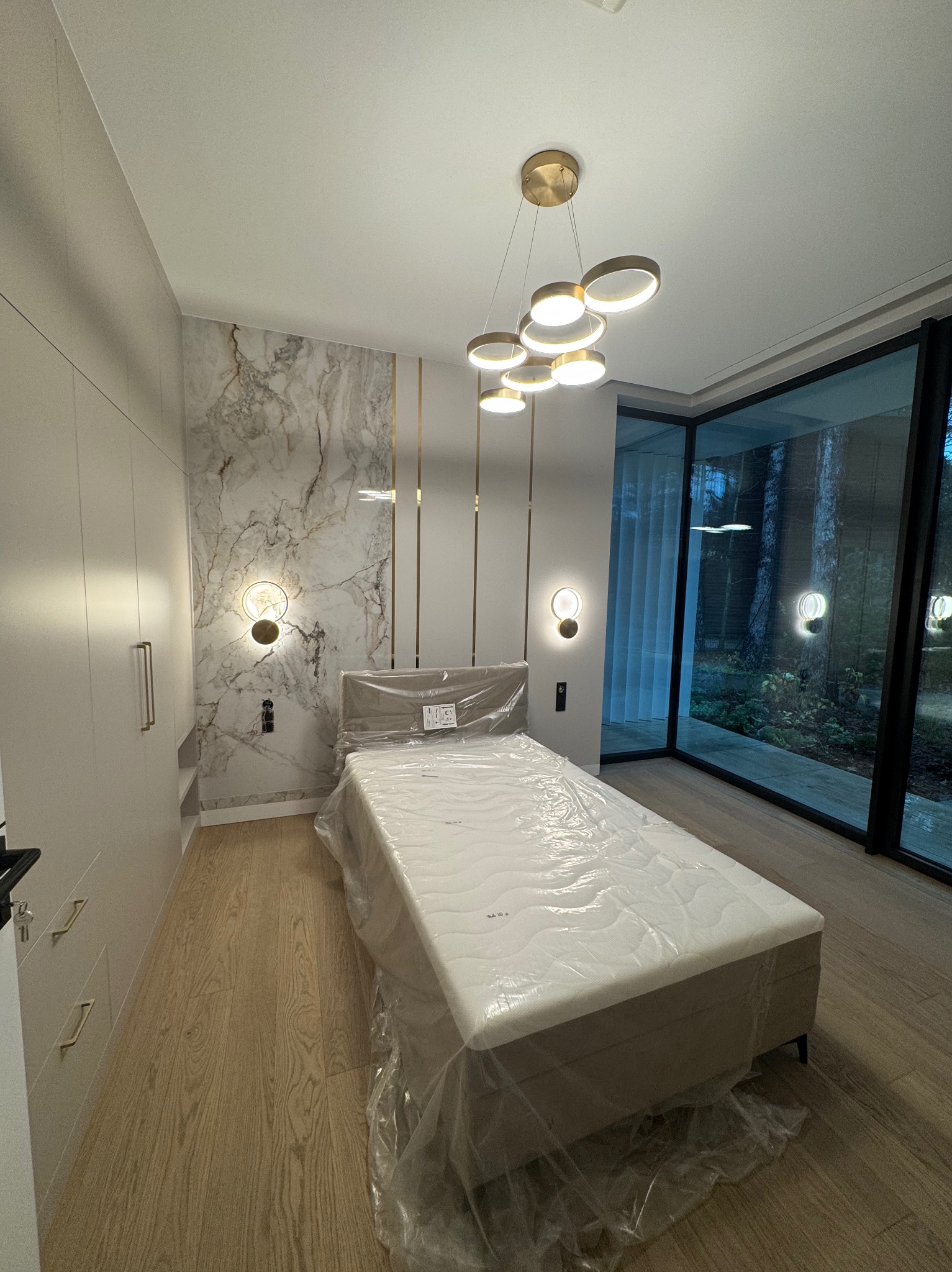 Nowoczesna sypialnia z marmurową ścianą, drewnianą podłogą i łóżkiem zabezpieczonym folią ochronną, oświetlona designerską lampą wiszącą i kinkietami.