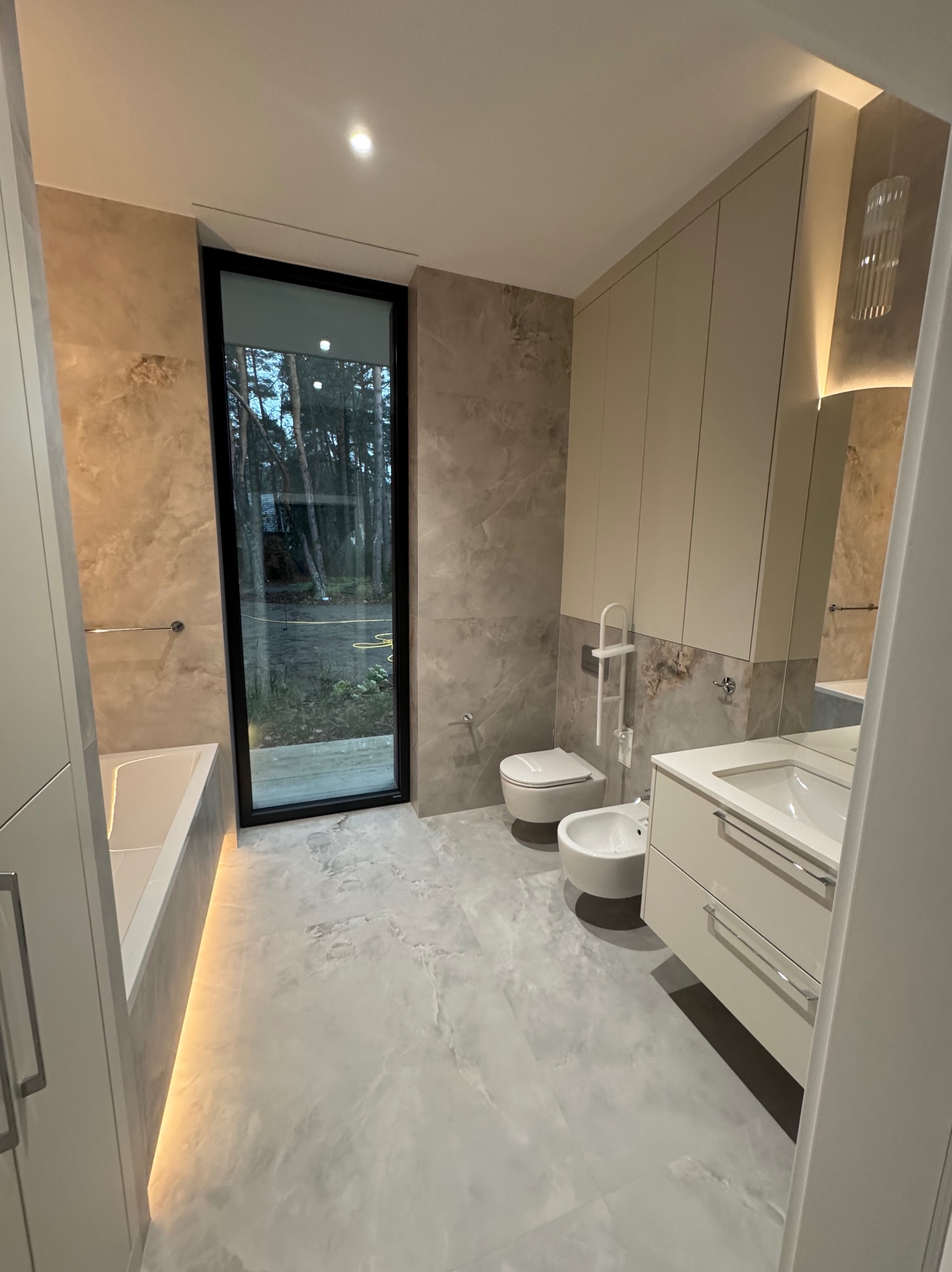 Nowoczesna łazienka z marmurowymi płytkami, podświetlaną wanną, dużym oknem z widokiem na zieleń, wiszącą toaletą i bidetem oraz szafkami w kolorze kremowym z oświetleniem LED.