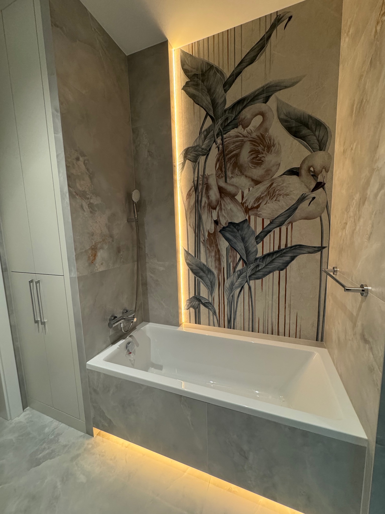 Wykończona łazienka z wanną obudowaną płytkami imitującymi marmur, oświetleniem LED pod wanną i na ścianie z motywem flamingów i roślin, oraz szafką w zabudowie.