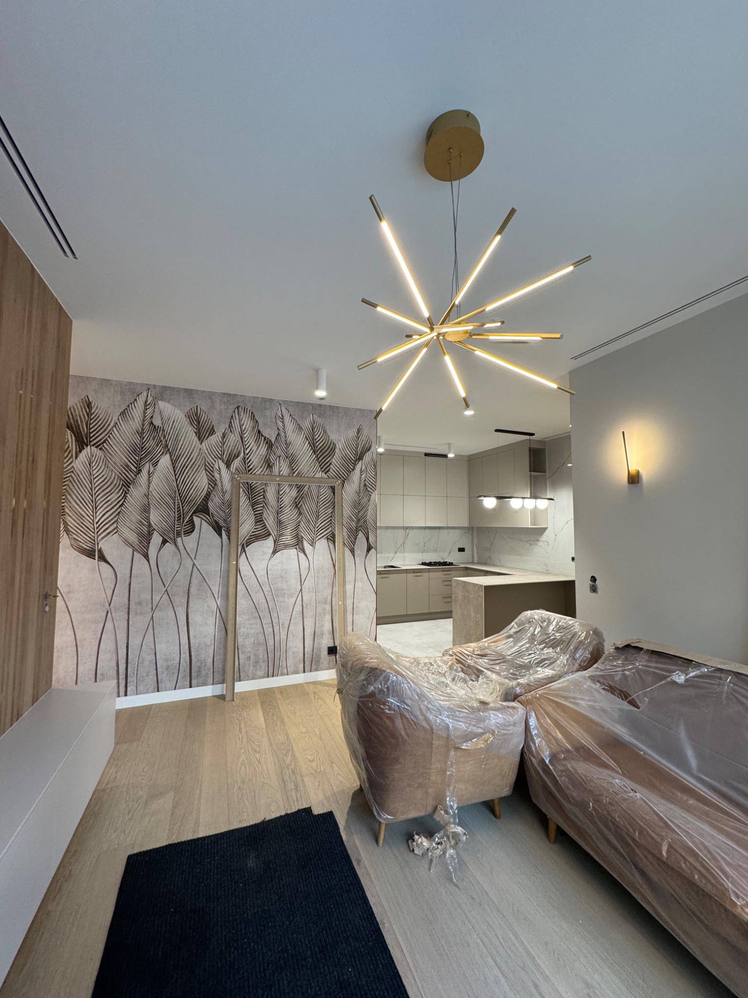 Salon w trakcie wykańczania: fotel i sofa zabezpieczone folią, na ścianie tapeta z motywem liści, nowoczesna lampa sufitowa i widok na kuchnię.