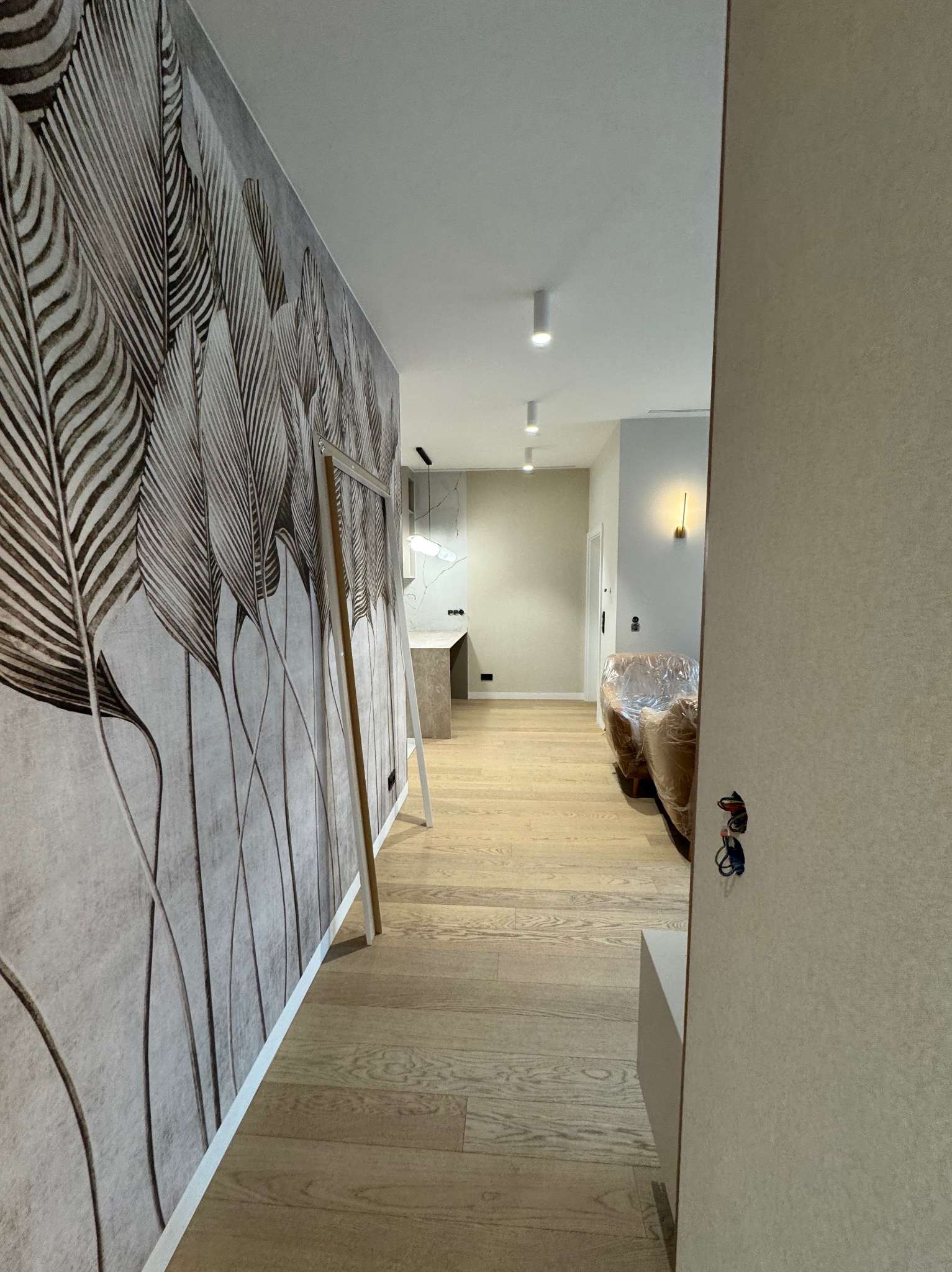 Wnętrze mieszkania w trakcie wykańczania: widoczna ściana z tapetą w motywie piór, drewniana podłoga, meble zabezpieczone folią, rama oparta o ścianę, widok na kuchnię i salon.