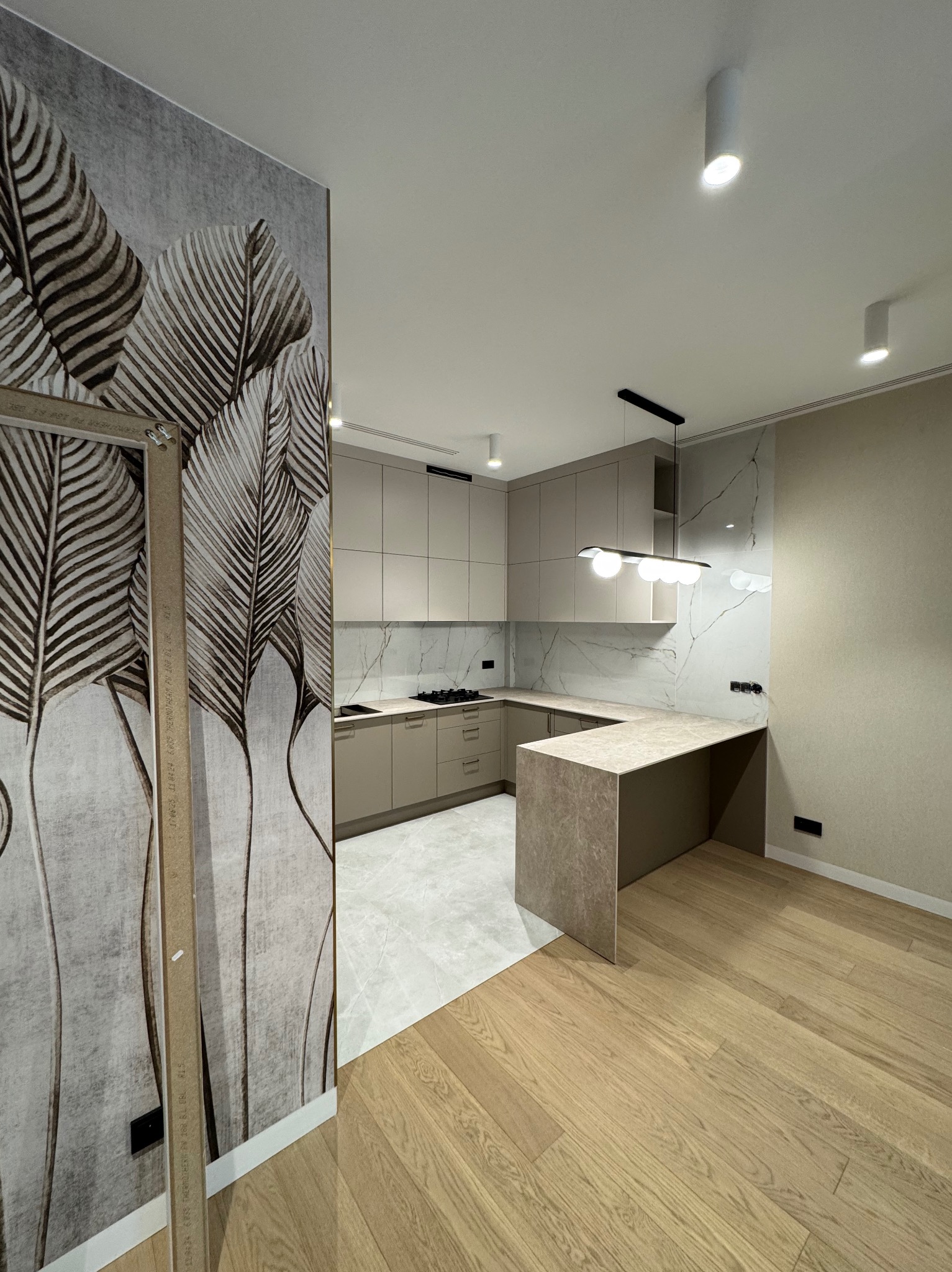 Nowoczesna kuchnia w beżach z marmurowym akcentem na ścianie i blacie, widok z perspektywy hallu z dekoracyjną tapetą w liście palmowe i drewnianą podłogą.