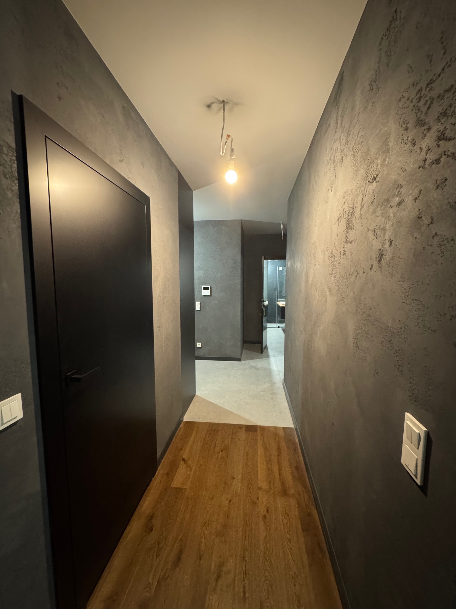 Korytarz po remoncie z drewnopodobną podłogą, ścianami pokrytymi dekoracyjną szarą farbą z efektem betonu i czarnymi drzwiami.
