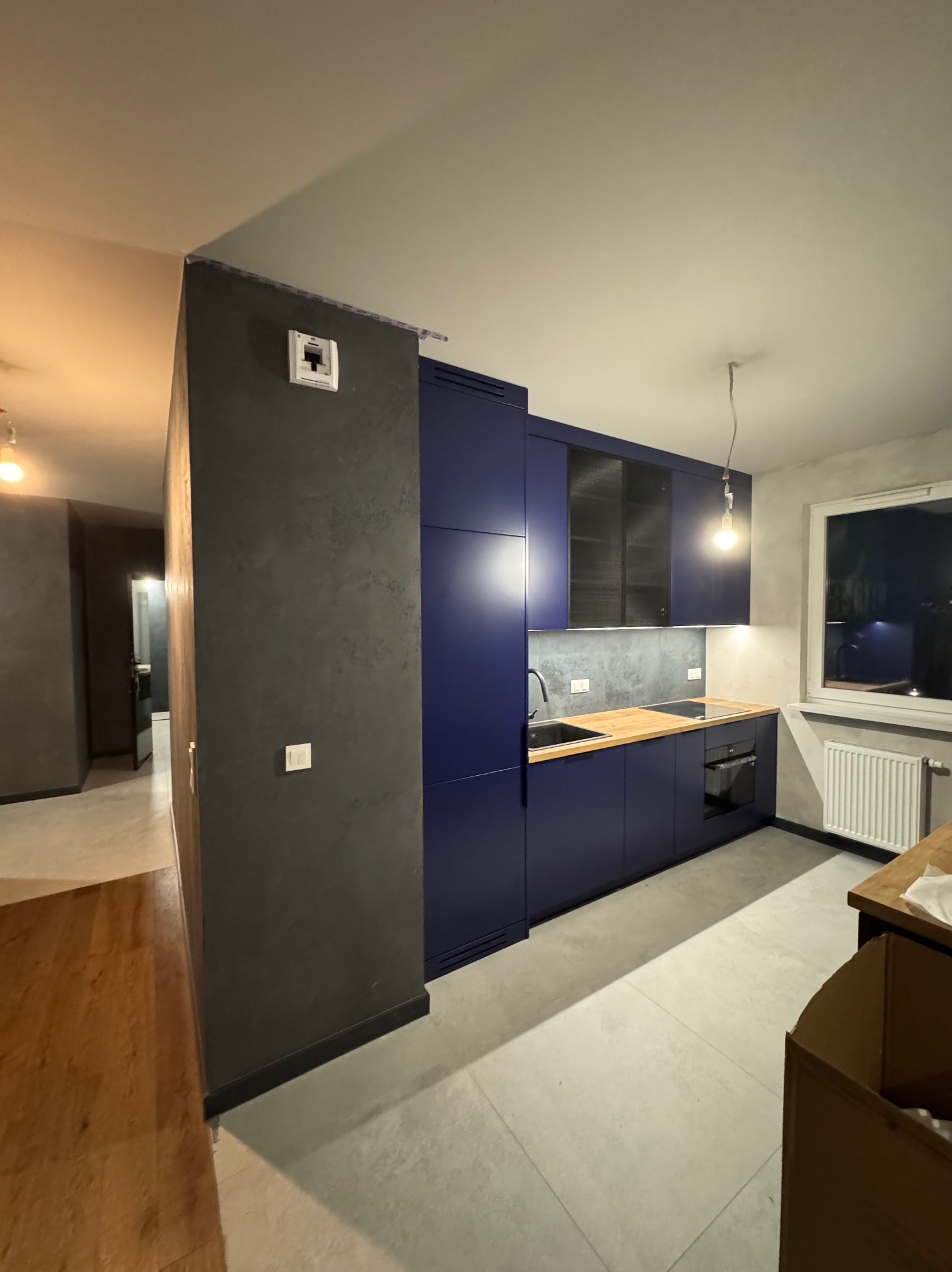 Nowoczesna kuchnia w odcieniach granatu z drewnianym blatem, szarą ścianą z efektem betonu i widoczną instalacją elektryczną sufitową przed montażem lampy.