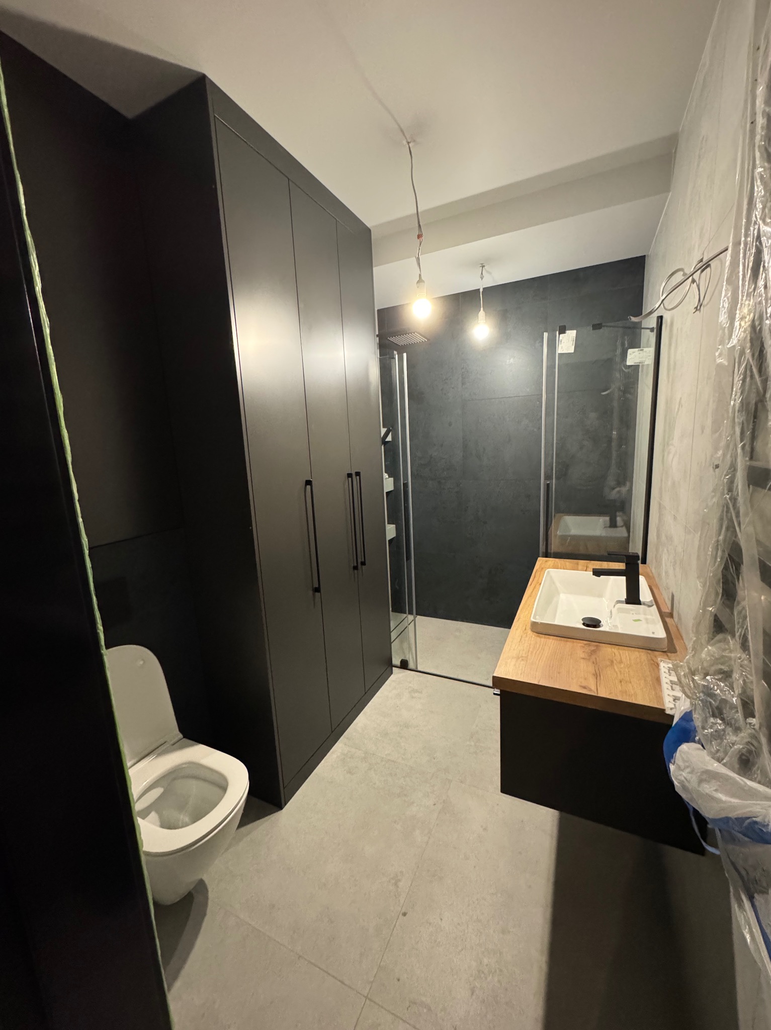 Nowoczesna łazienka z czarnymi szafkami, kabiną prysznicową z ciemnymi płytkami i umywalką na drewnianym blacie, widoczny także biały sedes.