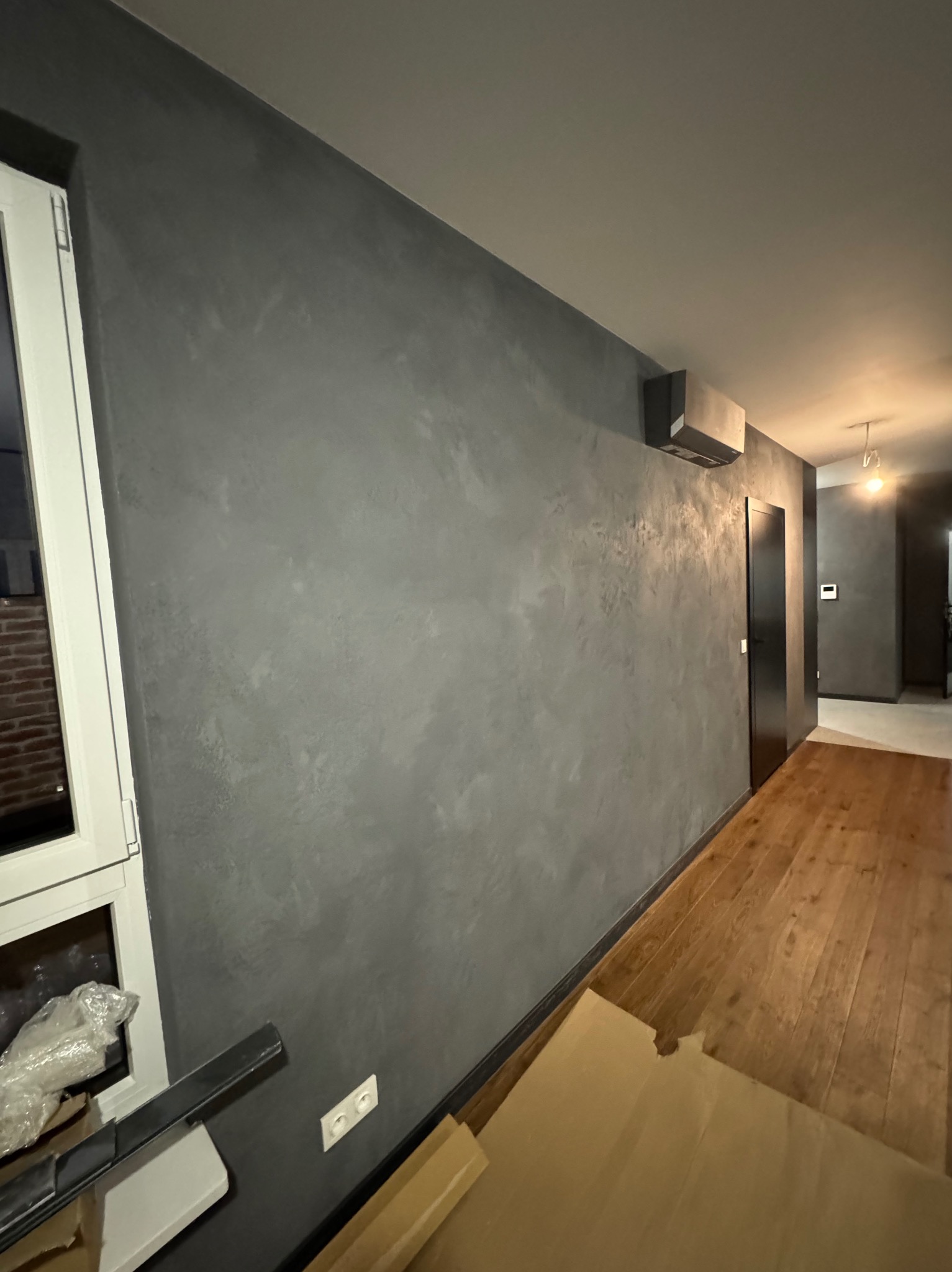 Wnętrze po wykonaniu gładzi dekoracyjnej w odcieniach szarości, widoczny fragment okna z parapetem, drewniana podłoga i czarne drzwi w głębi pomieszczenia.