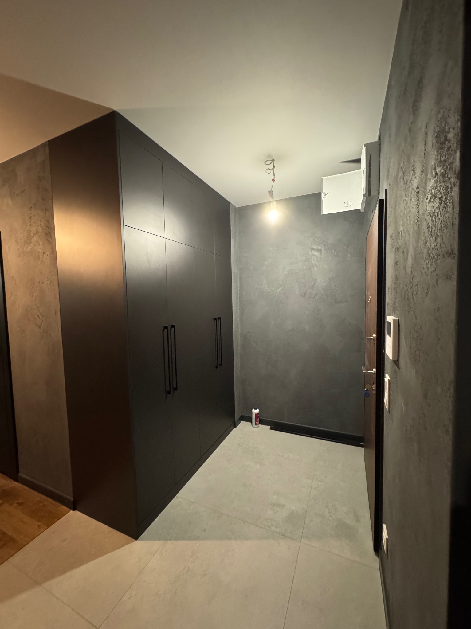 Wnętrze korytarza z dużą, matową, czarną szafą w zabudowie, szarą ścianą z efektem dekoracyjnym, szarą podłogą z płytek i częściowo widoczną drewnianą podłogą w innym pomieszczeniu.