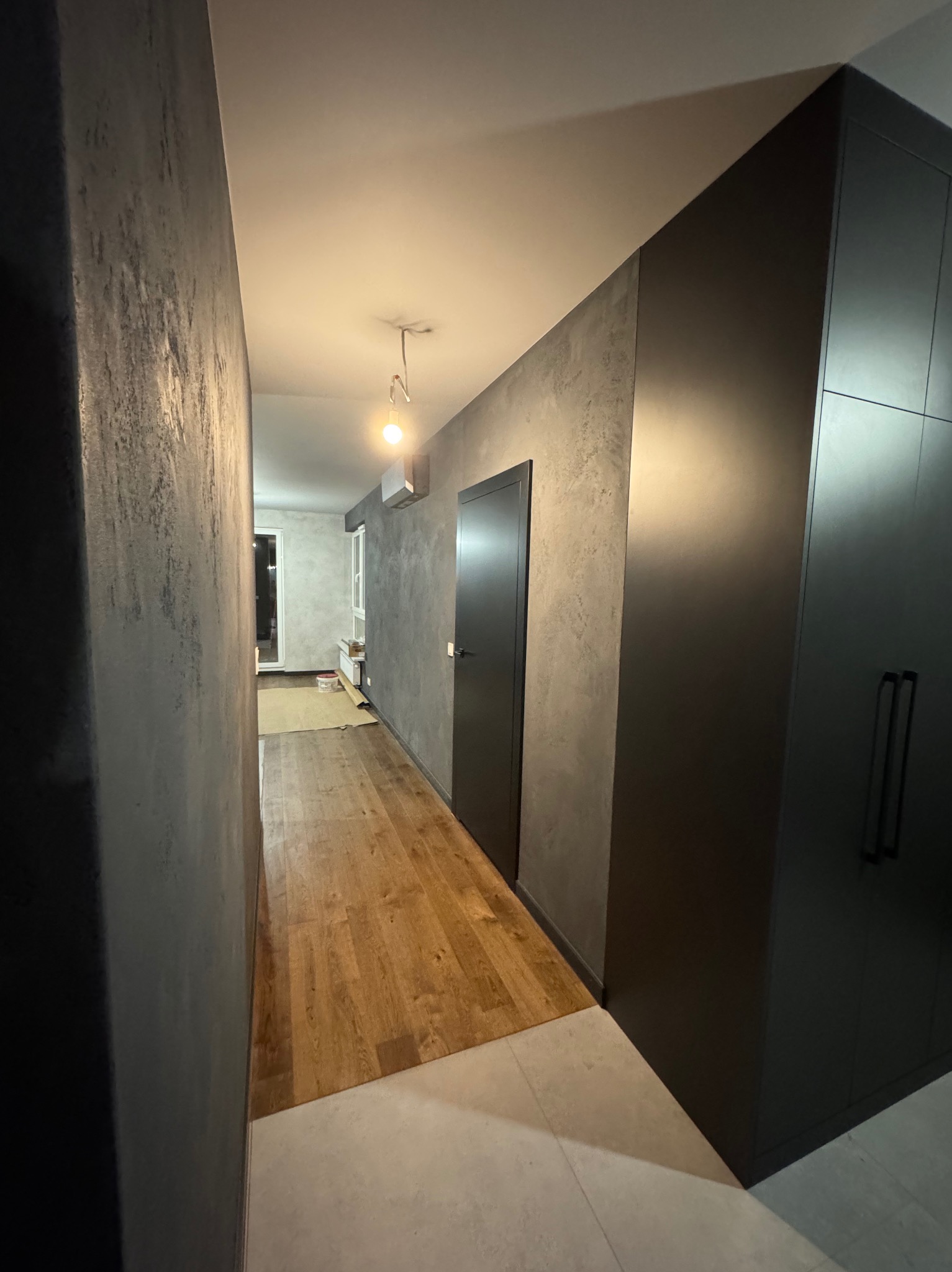 Korytarz po remoncie z drewnianą podłogą, ścianami w odcieniach szarości z efektem dekoracyjnym i zabudową meblową w kolorze grafitowym.