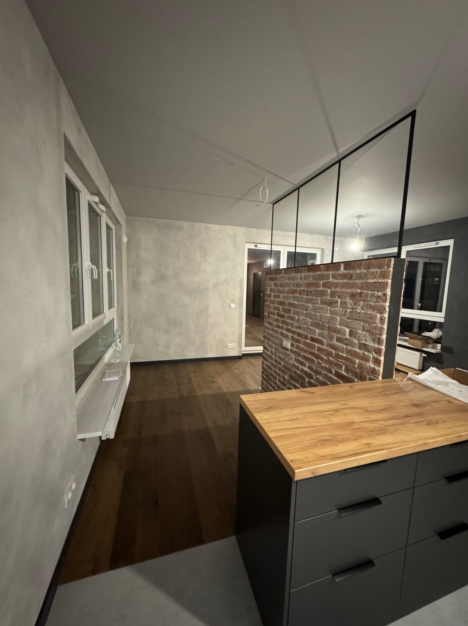Surowe wykończenie wnętrza w Warszawie: szara ściana z efektem betonu, drewniana podłoga, ceglana ścianka działowa z czarną ramą, widok na dalszą część mieszkania.