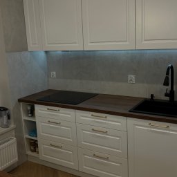 Rafał Stankiewicz - Jasna kuchnia na wymiar z drewnianym blatem i złotymi uchwytami, płyta indukcyjna i czarny zlew. Podświetlenie LED pod szafkami górnymi. Parkiet w jodełkę na podłodze.