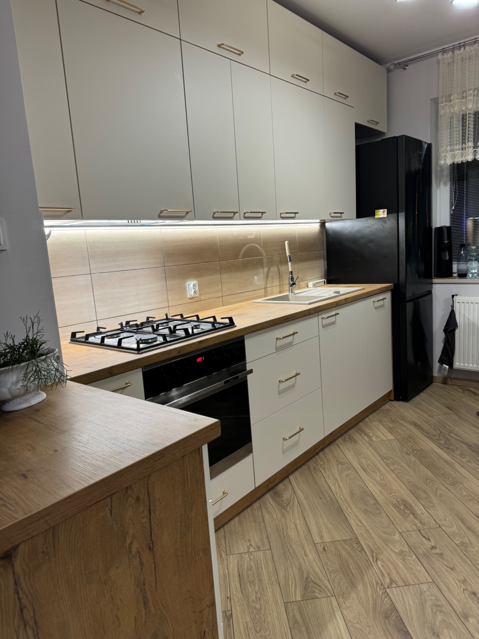Jasna kuchnia z drewnianymi akcentami: zabudowa z szafkami górnymi i dolnymi, płyta gazowa, piekarnik, zlew i czarna lodówka. Uwagę przykuwają złote uchwyty.
