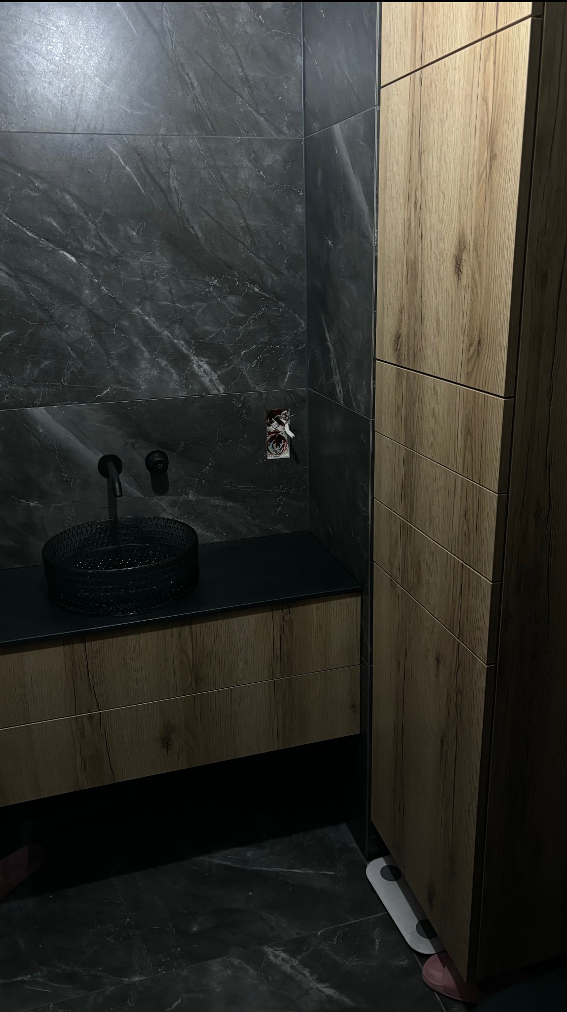 Nowoczesna łazienka: czarna umywalka na blacie z drewnopodobną szafką i marmurową ścianą. Obok wysoka szafa z frontami w odcieniach drewna. Minimalistyczny design.