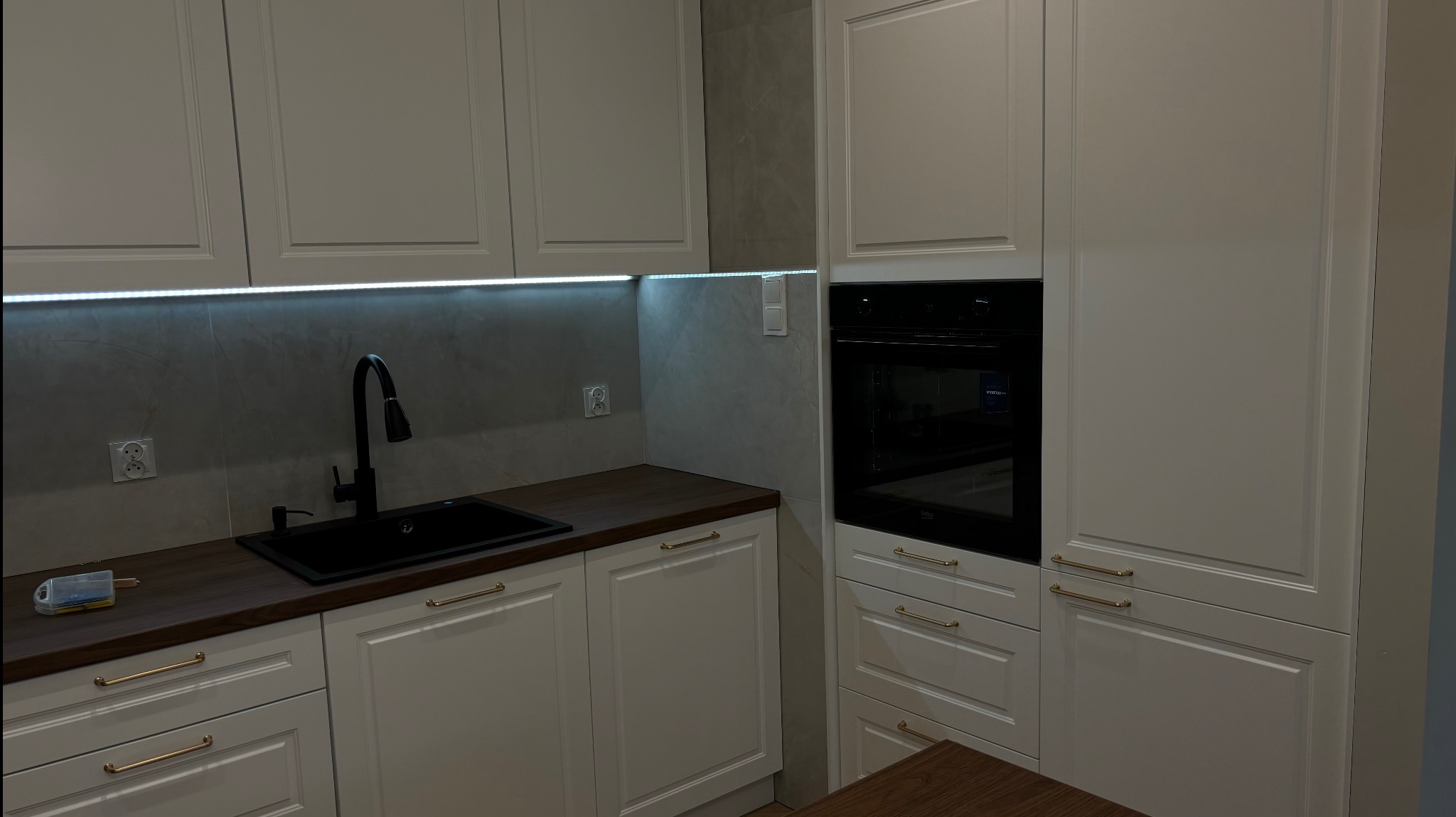 Jasna, klasyczna kuchnia z ciemnym blatem i czarną armaturą. Oświetlenie LED podszafkowe podkreśla subtelny wzór płytek ściennych.